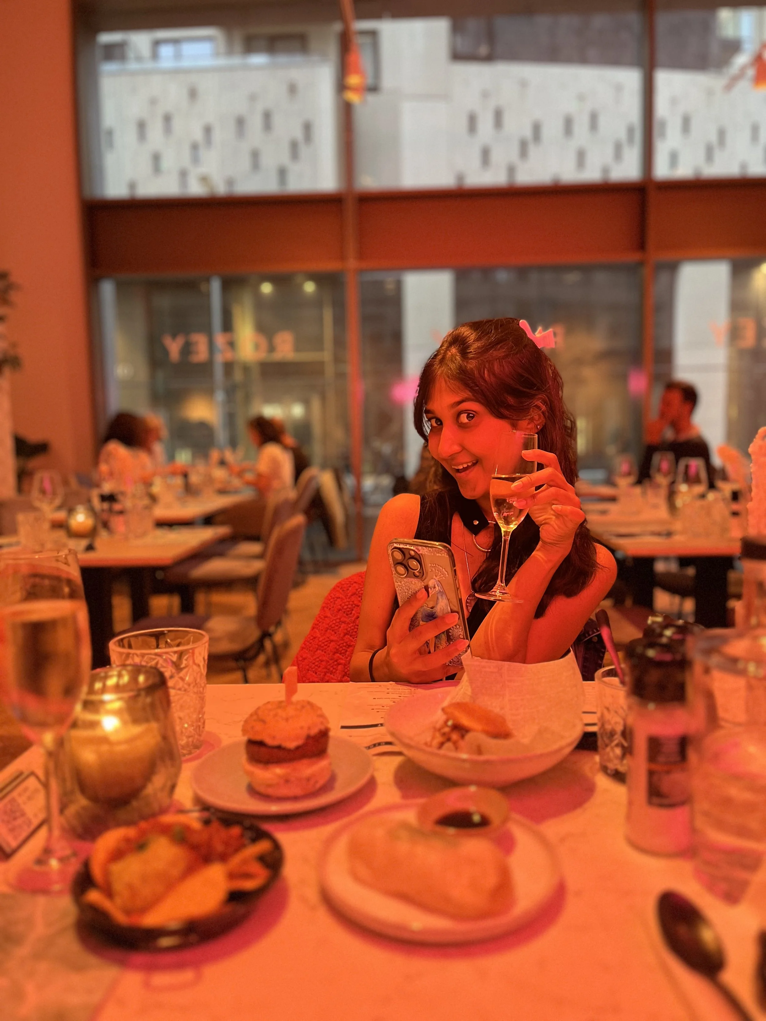 A woman smiling and holding a glass of champagne in a restaurant with a table of desserts in the foreground.