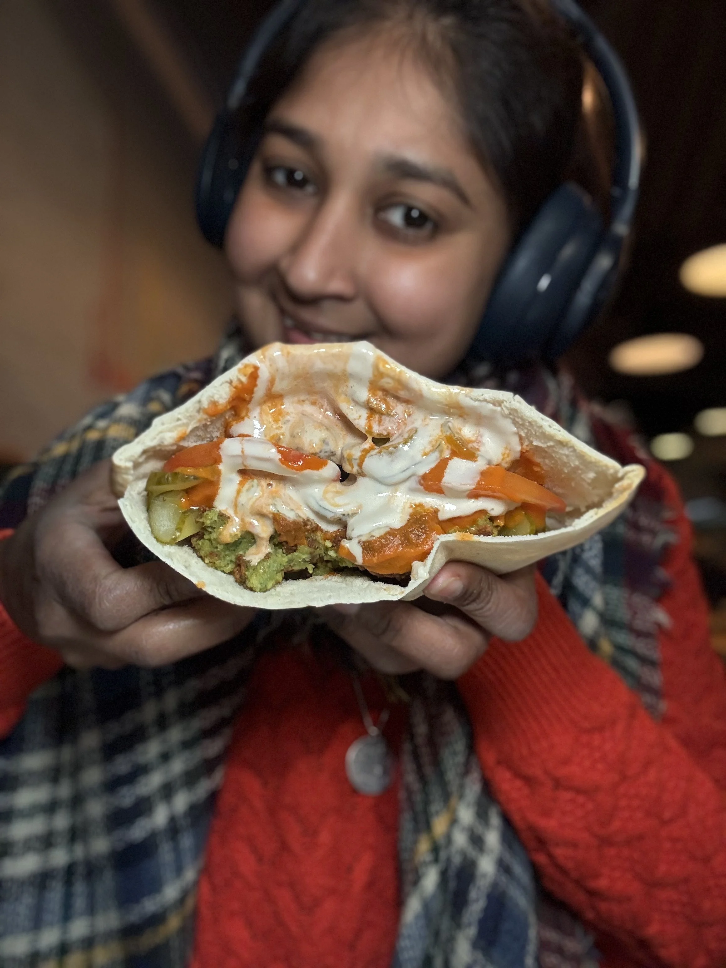 A woman with black hair wearing headphones holds a taco with various fillings including avocado, sour cream, and vegetables close to the camera.