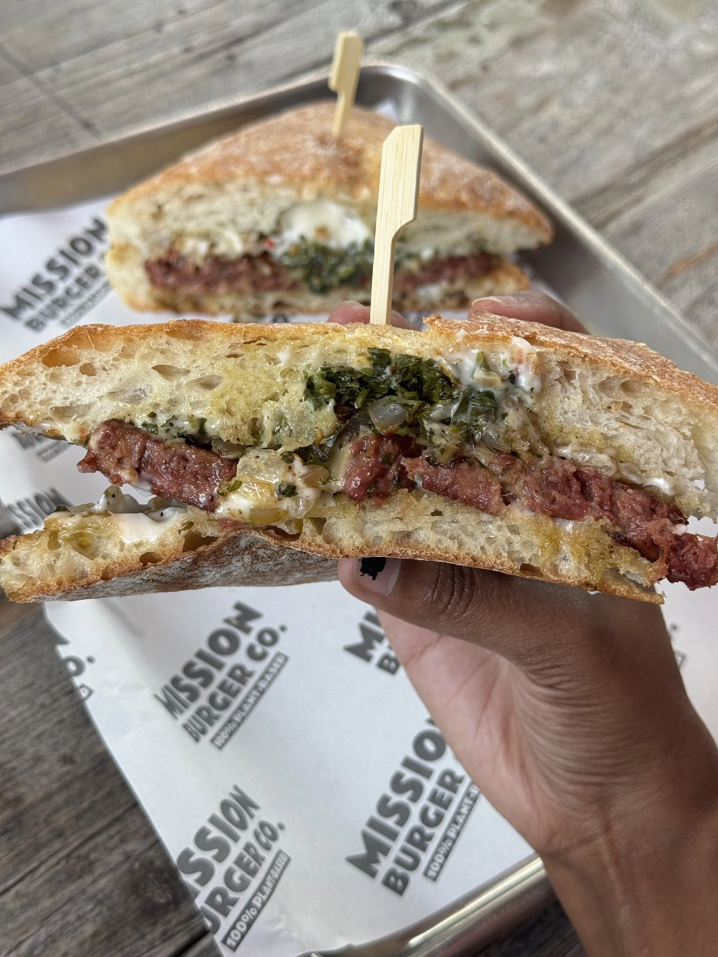 A hand holding a sliced meat sandwich with lettuce, mayonnaise, and cheese, on a toasted hoagie bun, with the rest of the sandwich visible in the background. The sandwich is on a tray with paper that says 'Missouri Burger' on it, and the tray is on a wooden surface.