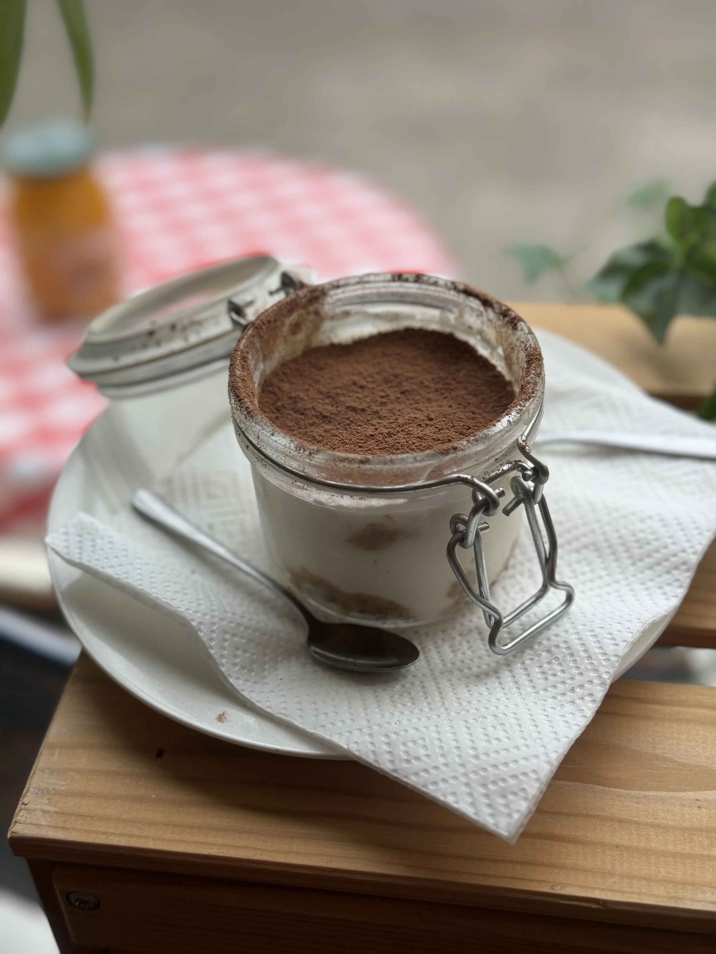 A jar of chocolate mousse topped with cocoa powder on a white plate with a napkin underneath, sitting on a wooden table with a spoon nearby, blurred background with green plants and a pink checkered cloth.