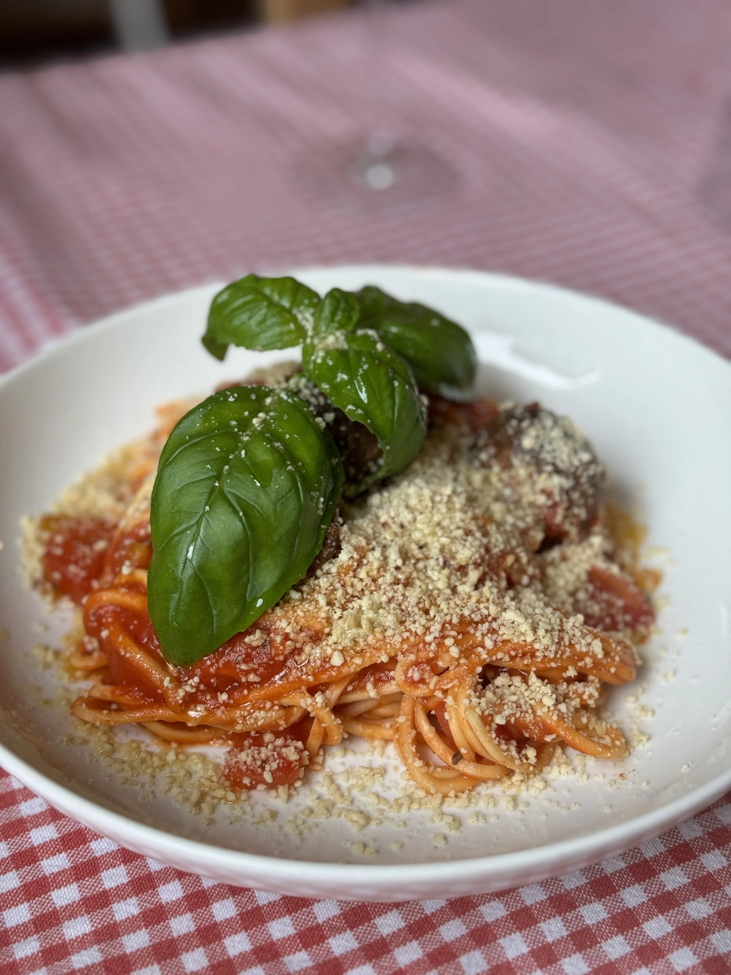 Spaghetti with tomato sauce, topped with fresh basil leaves and grated cheese, served in a white bowl on a red and white checkered tablecloth.