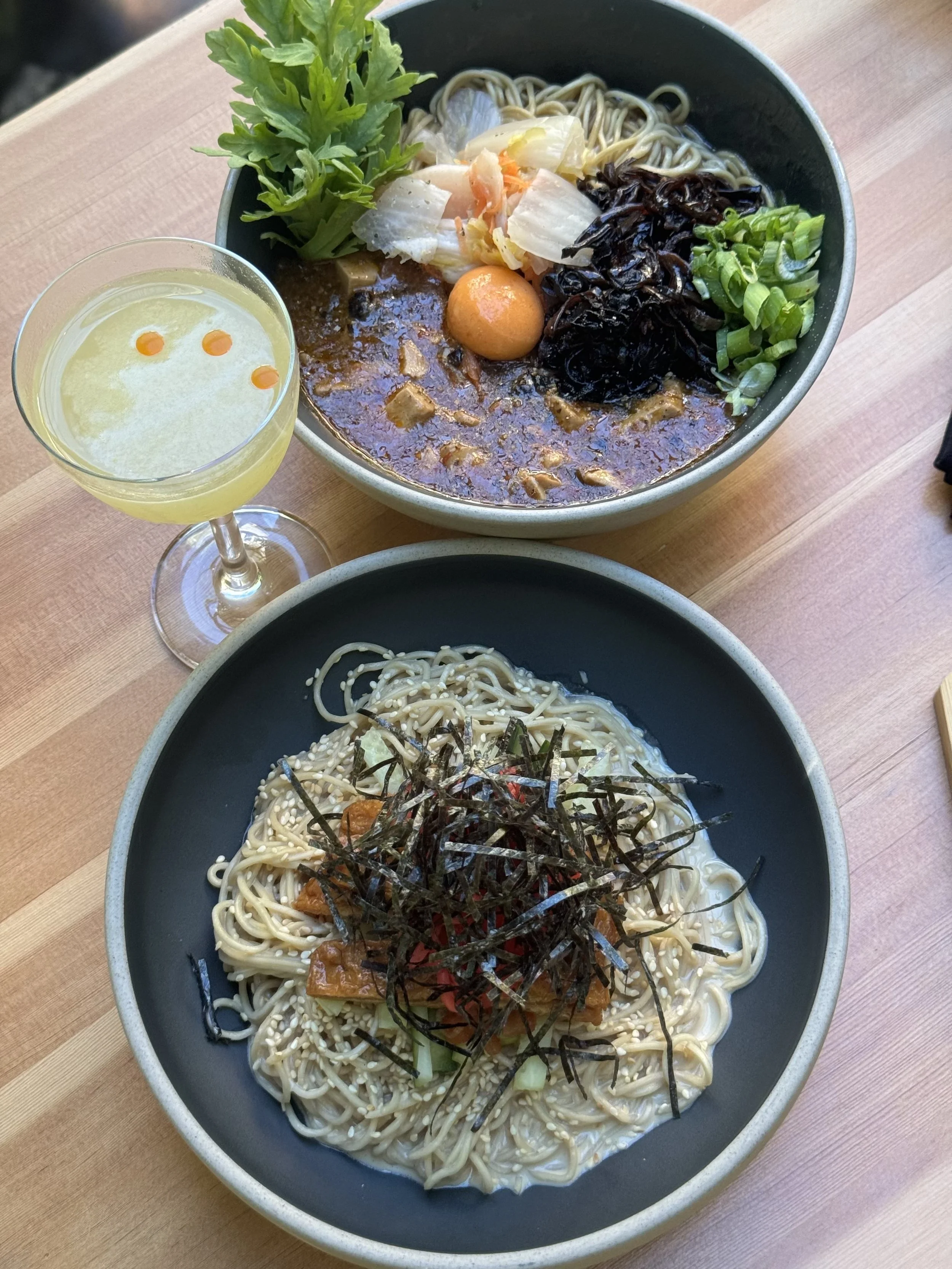 A bowl of ramen noodles topped with seaweed, green onions, and seasonings, with a side of a soft-boiled egg and vegetables, accompanied by a glass of white wine with three drops of hot sauce.