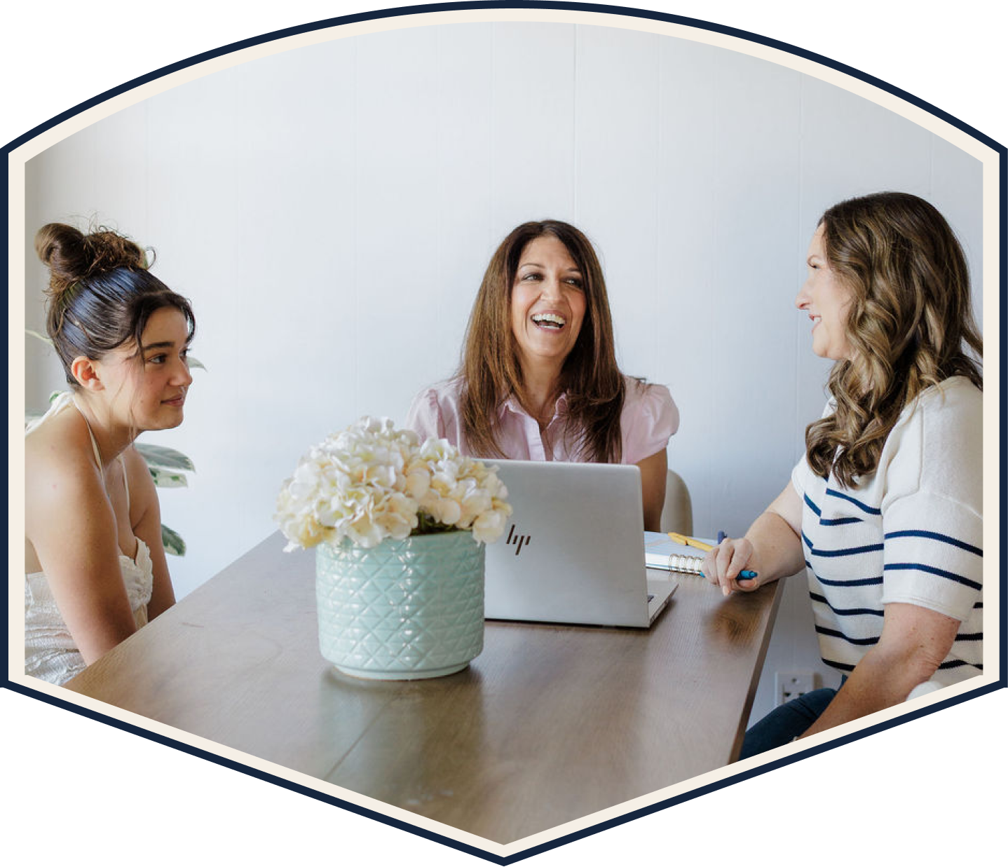 Three women are sitting at a table having a conversation about college admissions, with a laptop, notebook, and pen in front of them. A flower arrangement is on the table, and they are smiling and engaging with each other.