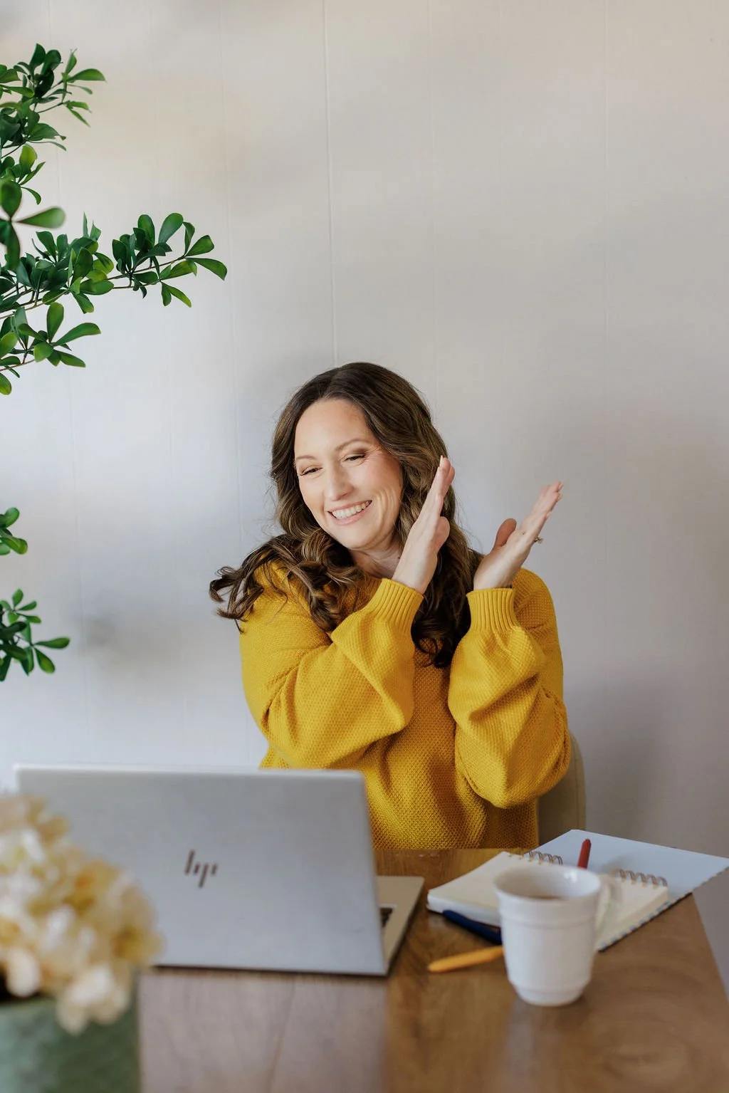 A woman with long brown hair, wearing a yellow sweater, sitting at a desk with a laptop, notebooks, and a coffee mug, smiling and cheering for a student she is helping. There is a potted plant on the left side of the scene.