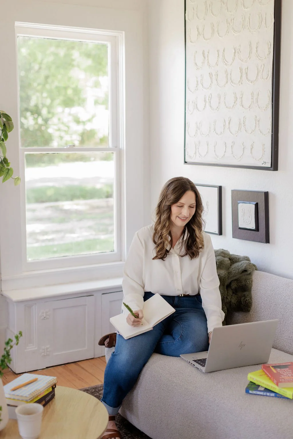 A woman sitting on a gray couch with a laptop and notebooks, smiling and writing in a notebook in a well-lit living room with framed artwork on the wall and a window showing greenery outside.