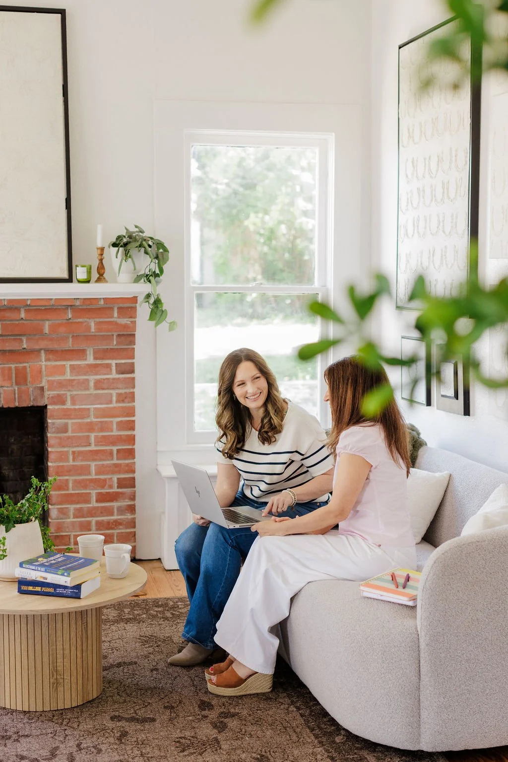 Two women sitting on a light gray sofa in a bright living room, smiling and talking while looking at a laptop. One woman has long wavy brown hair and wears a white and navy striped shirt, the other has long brown hair and wears a pink top. A small wooden coffee table with books, cups, and a plant is in front of them. The room has a brick fireplace, large window, and minimal wall art.