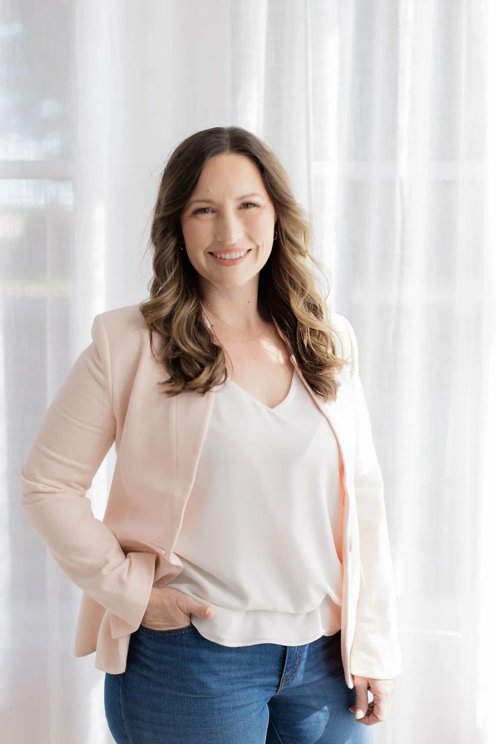 A woman with long, wavy brown hair smiling at the camera, wearing a light pink blazer, white blouse, and blue jeans, standing in front of a sheer white curtain