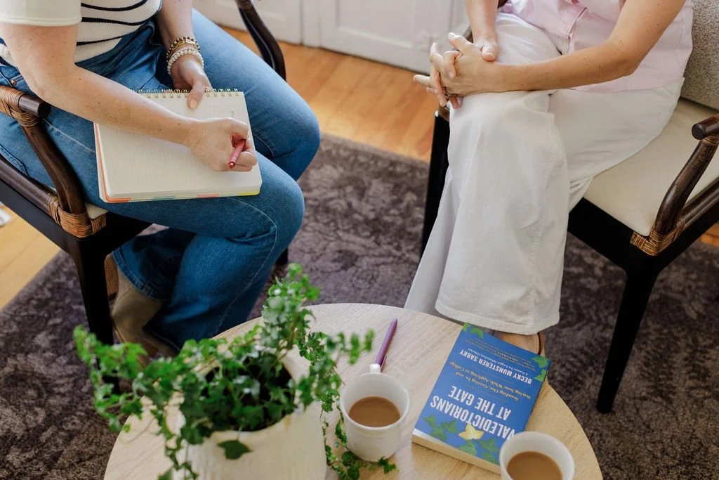 Two women sitting in chairs having a conversation; one taking notes with a notepad and pen, the other with hands clasped, wearing a pink shirt and white skirt; small round table with potted plant, notebooks, and coffee cups between them.