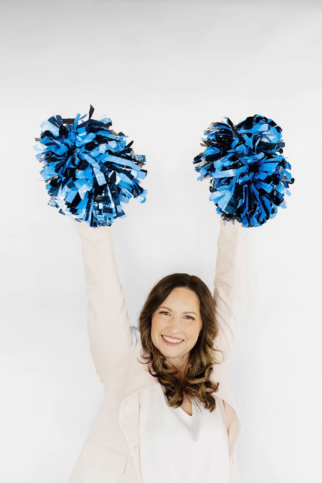 Woman in a light-colored blazer holding up blue pom-poms, smiling against a plain white background.