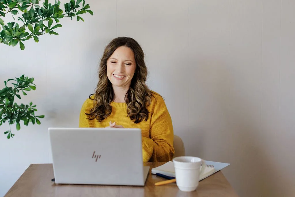 A woman with long brown hair, wearing a yellow sweater, smiling and working on a white laptop at a wooden desk, with a white coffee mug and a notebook nearby, next to a green plant, against a plain gray wall.