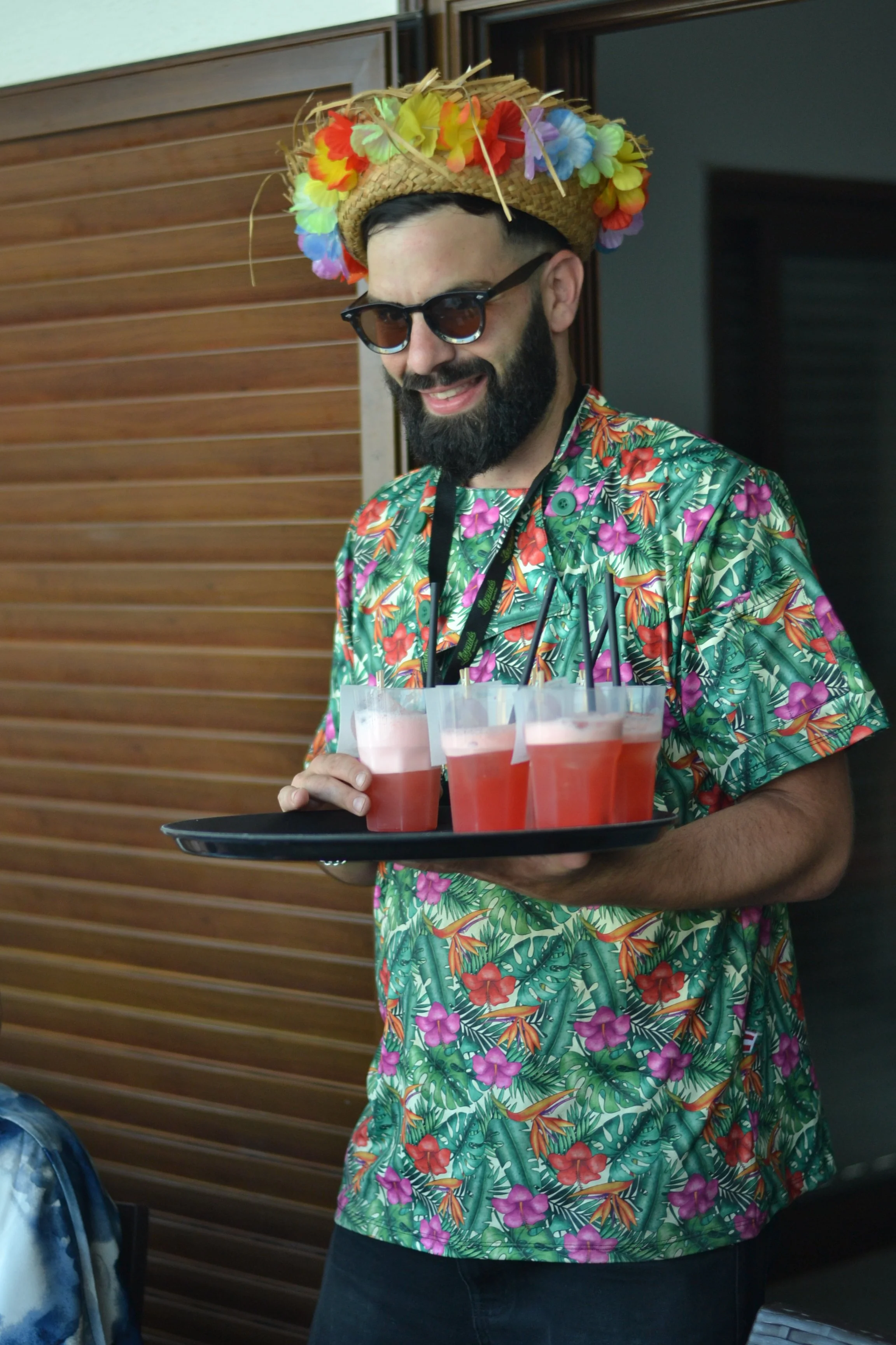 Homem de camiseta florida, usando óculos escuros, chapéu de palha decorado com flores coloridas, segurando um bandeja com copos de drink rosa com gelo e canudos pretos, em ambiente com parede de madeira ao fundo.