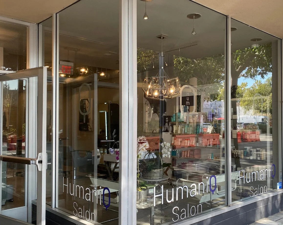 View of a salon named HumaniQ Salon through large glass windows and door, with hair and beauty products visible inside and trees outside.