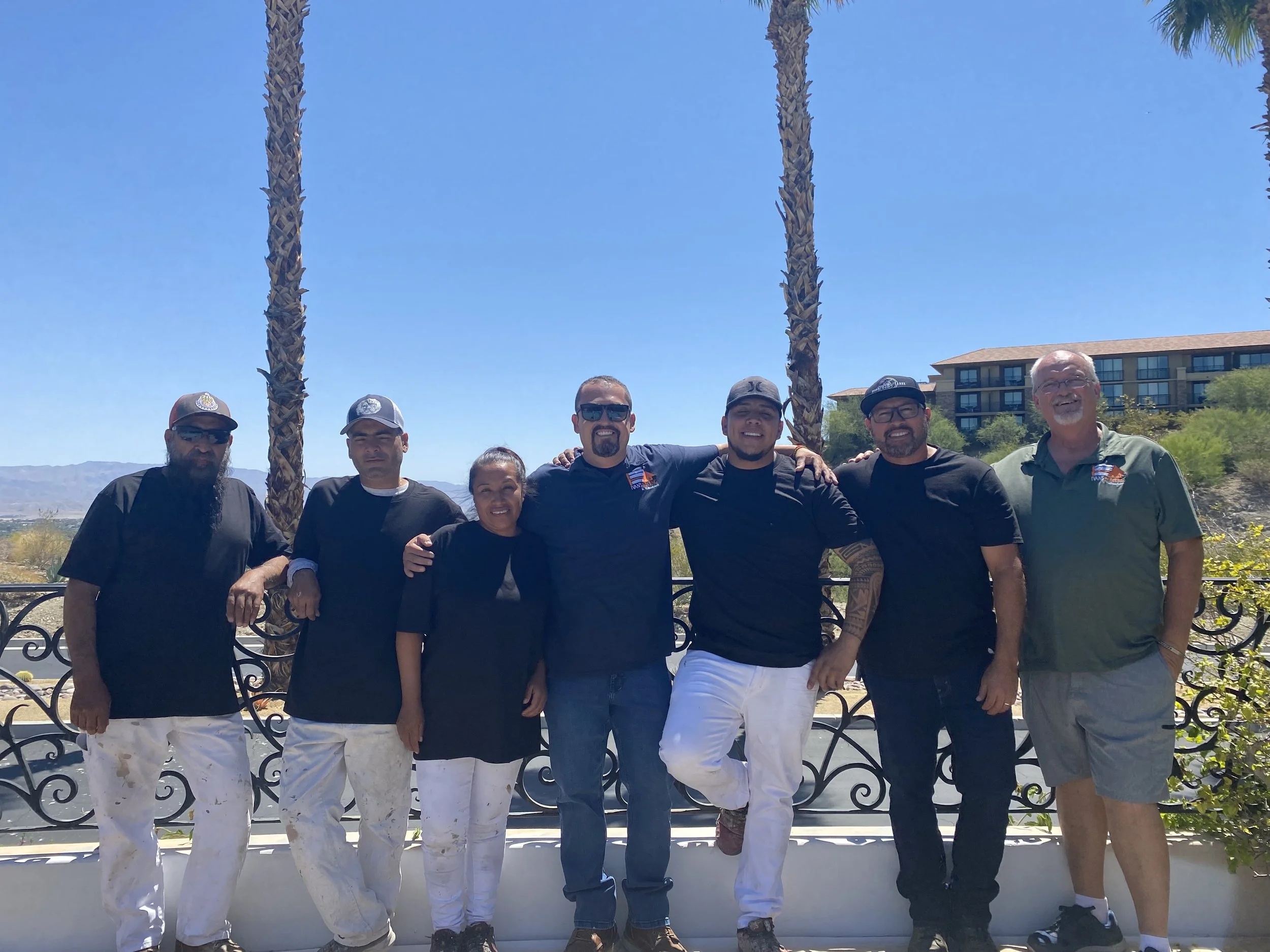 Group of seven people standing outdoors near a black decorative fence with two tall palm trees behind them, blue sky, and a building in the background. The group appears to be smiling and posing for a photo.