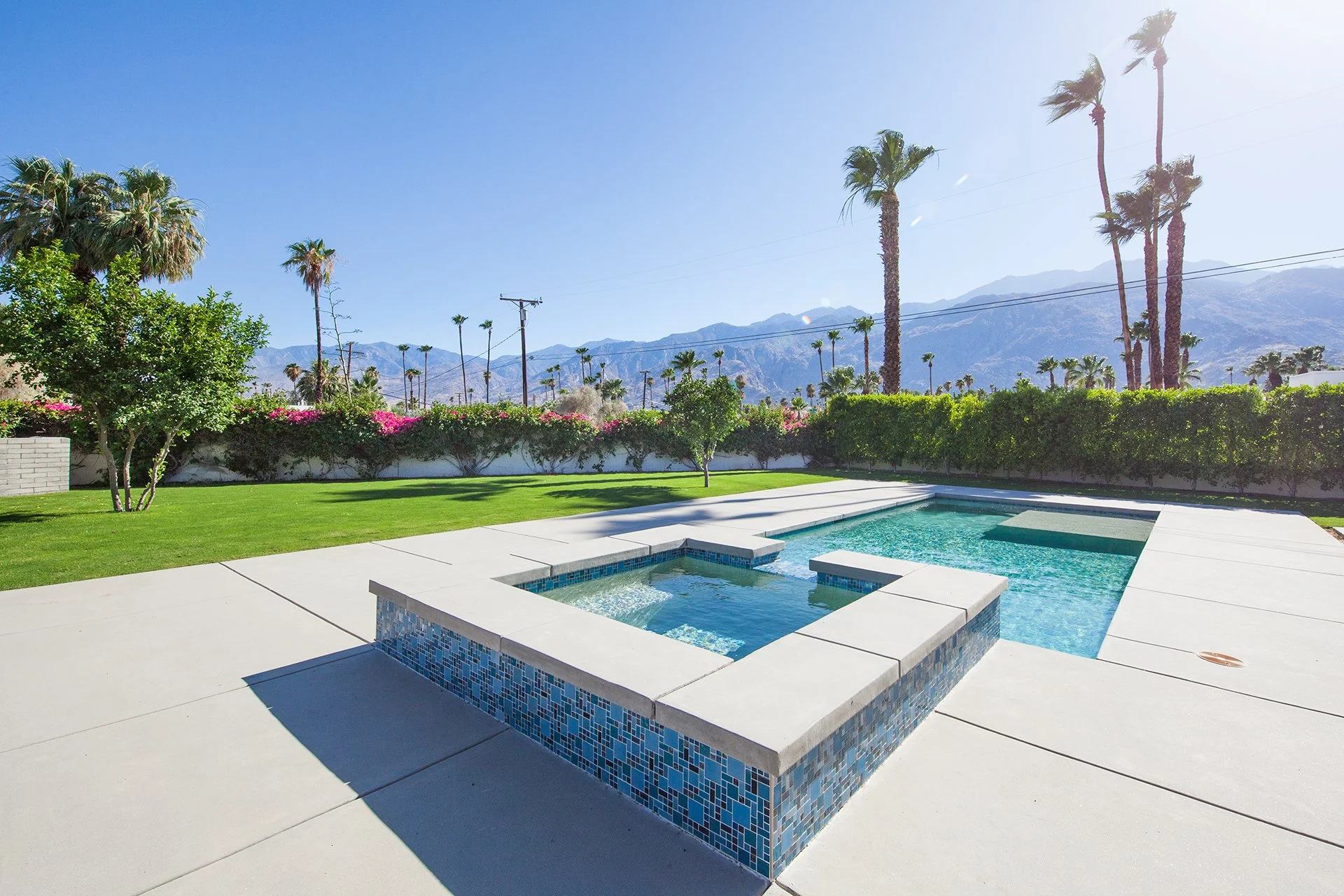 Backyard with swimming pool and hot tub, green lawn, trees, bushes, and mountain view under a clear blue sky.