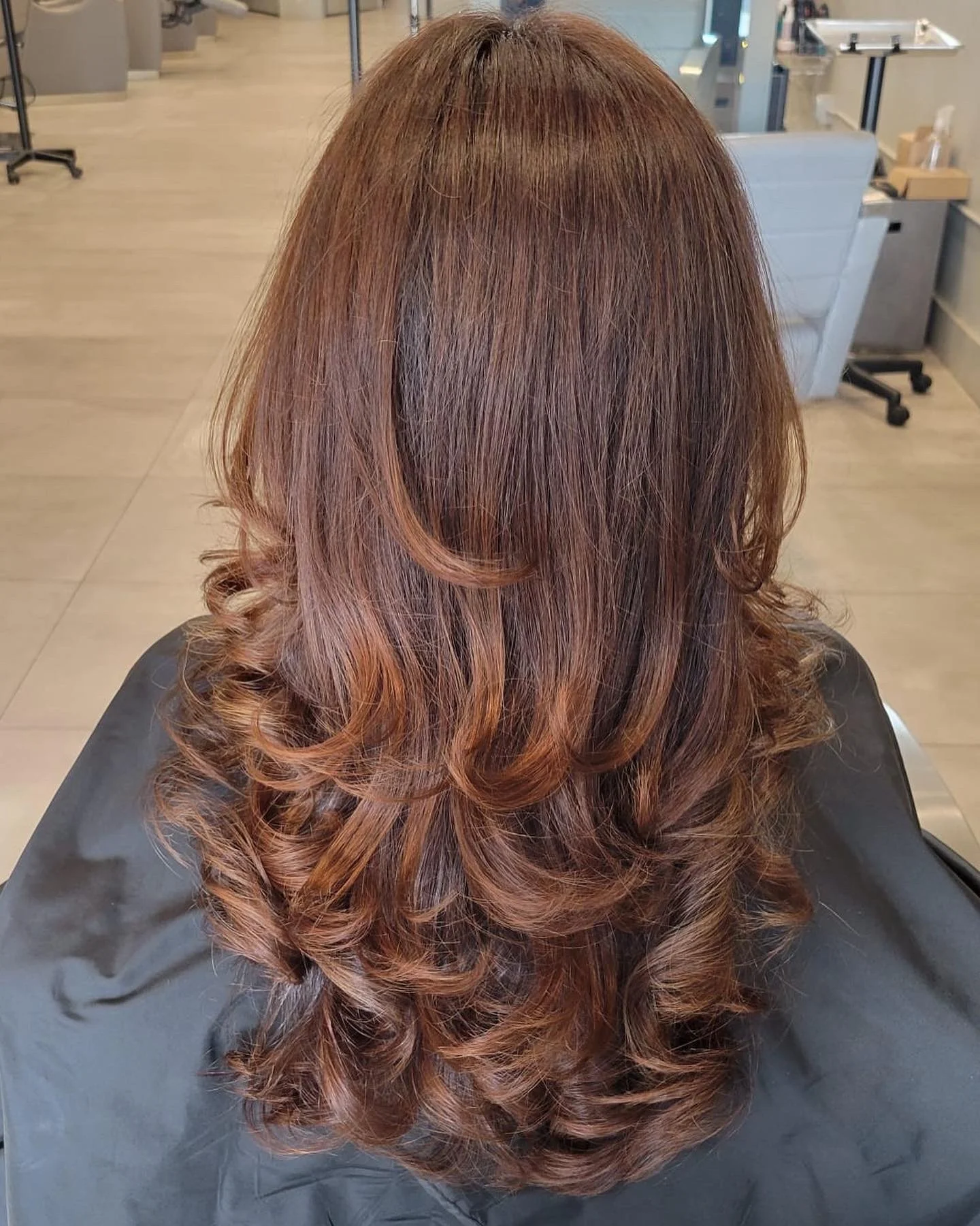 The image shows the back of a person's head with long, layered, reddish-brown hair styled in loose curls, seated in a salon chair with a hairdressing station visible in the background.