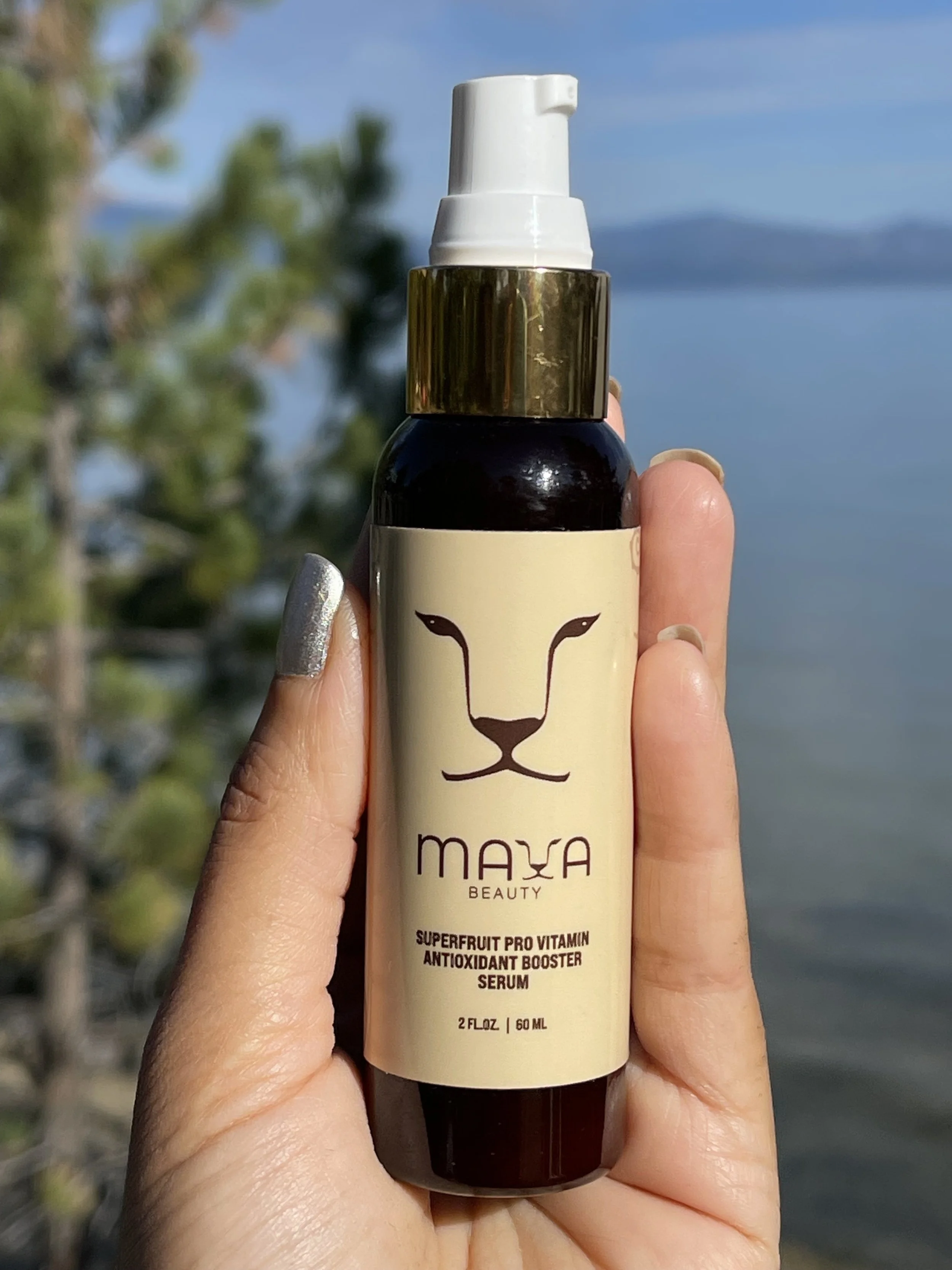A hand holding a bottle of Maxa Beauty Superfruit Pro Vitamin Antioxidant Booster Serum outdoors with a blue sky and water background.
