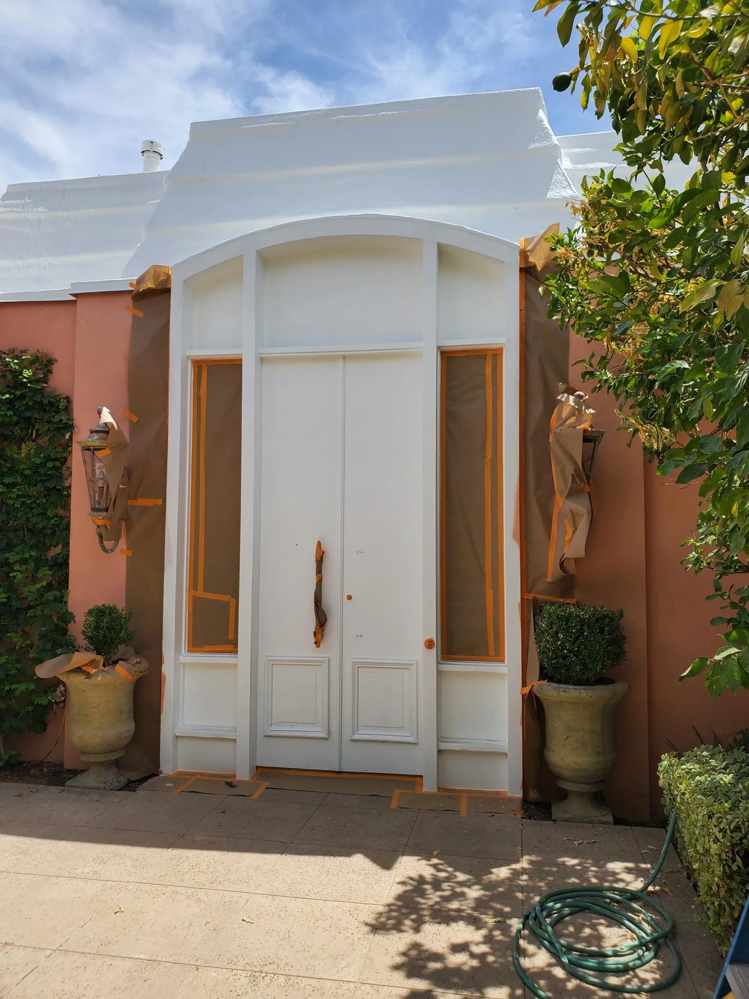 A house with a large white wooden door and glass windows is under renovation, with masking tape and paper covering parts of it. There are plants in large pots on either side, and a green garden hose on the ground. The sky is blue with some clouds.