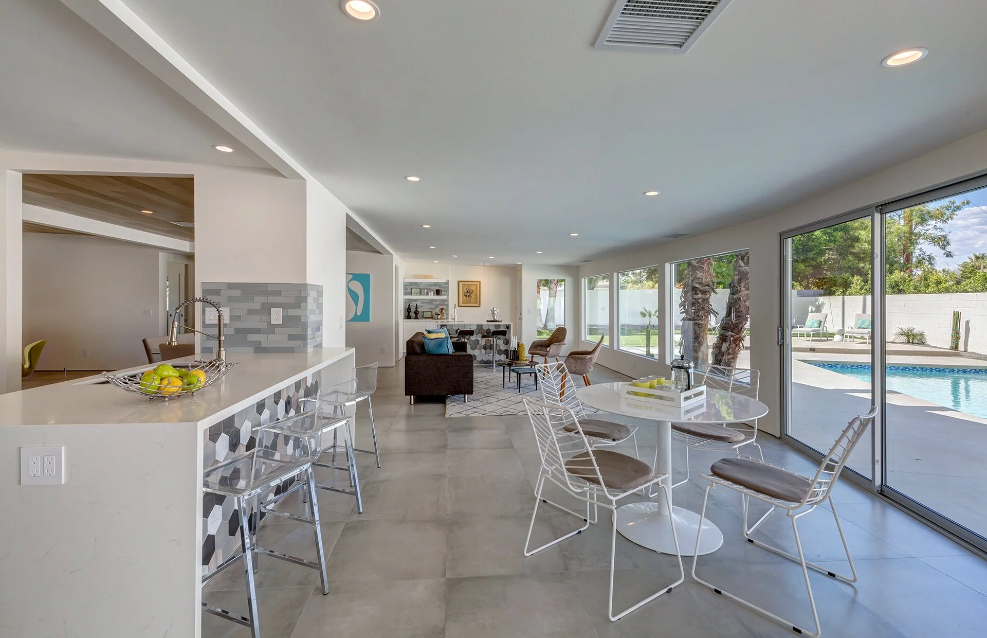 Bright, open living room with large windows and sliding glass doors overlooking a backyard pool, furnished with modern chairs, a round dining table, and a couch.