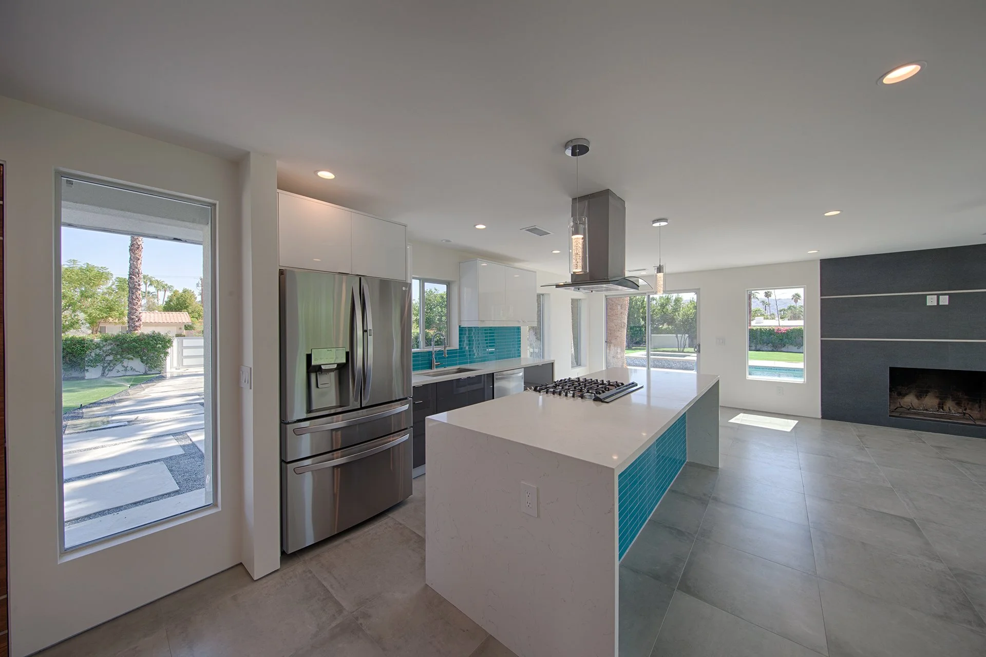 Modern kitchen with white countertops, stainless steel refrigerator, blue tiled backsplash, and large windows showing greenery outside.