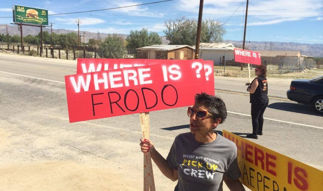 Two women holding signs on a roadside in a desert landscape. The signs read 'WHERE IS ? FRODO' and 'WHERE IS BUDDY'. The background features mountains, a McDonald's billboard, and a few small buildings.