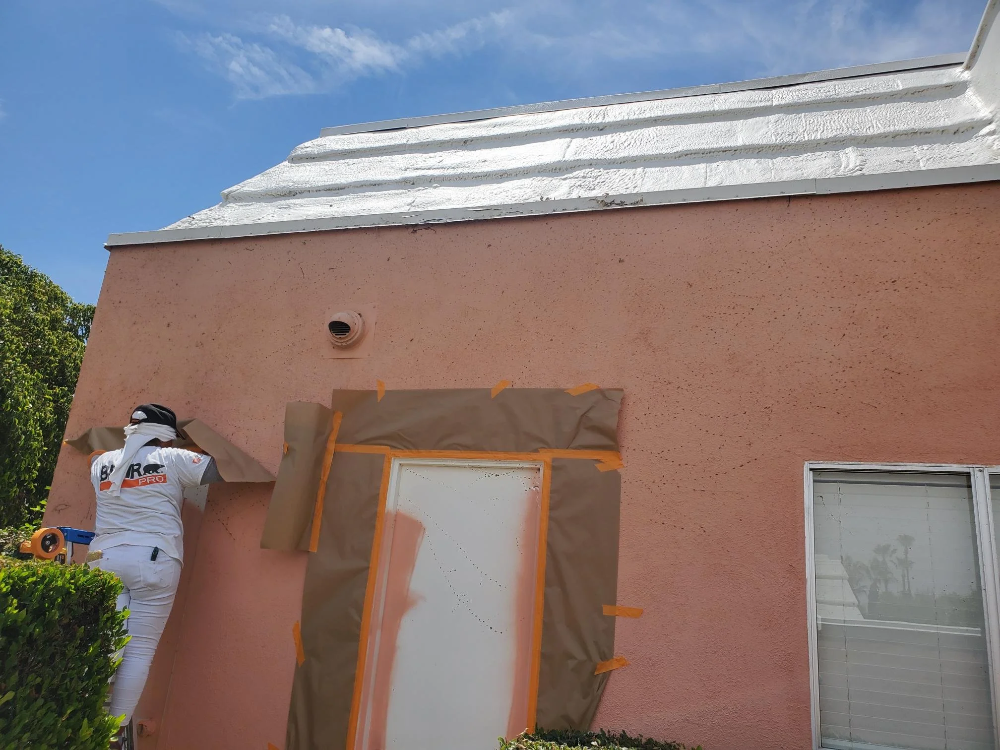 A person wearing white clothes and a cap is spray painting the wall of a house. The house has a peach-colored exterior wall and white trimming on the roof. The window has rectangular white grids, and there is a vent on the wall. The person is working with tape and paper to cover areas during painting, and there are trees and a blue sky in the background.