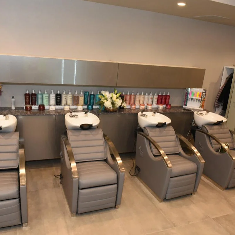 A modern salon with three reclining chairs positioned under a mirror, each with a sink for hair washing at head level, and a countertop behind with various hair care products, a flower arrangement, and hair color samples.