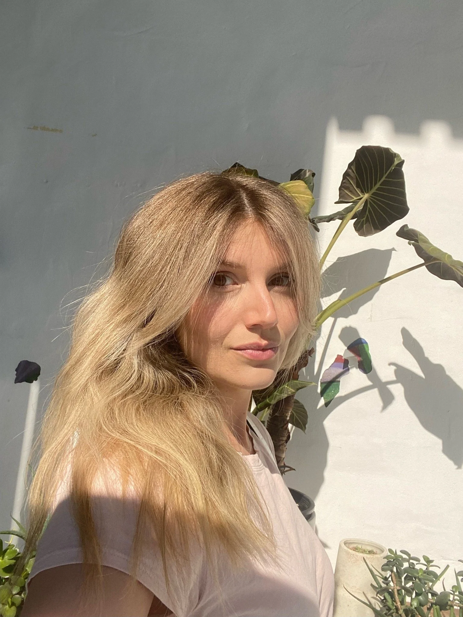 A woman with long blonde hair and a light pink shirt standing next to potted plants with large leaves, bright sunlight casting shadows on the wall behind her.