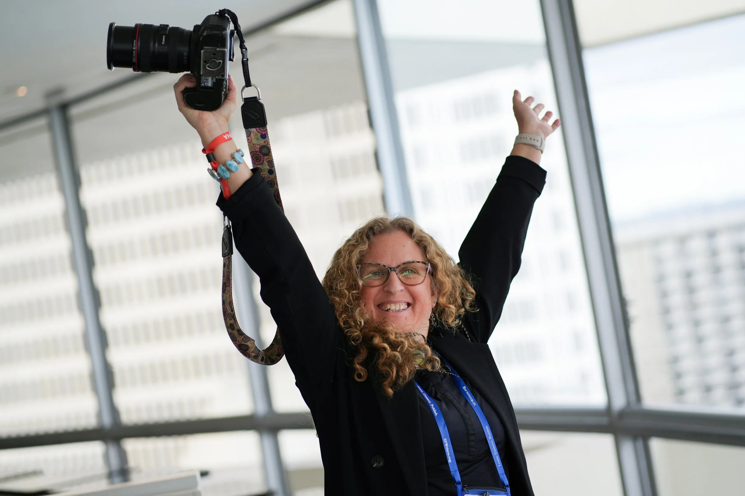 A woman with glasses smiling and raising her arms, holding a camera in one hand, inside a building with large windows.