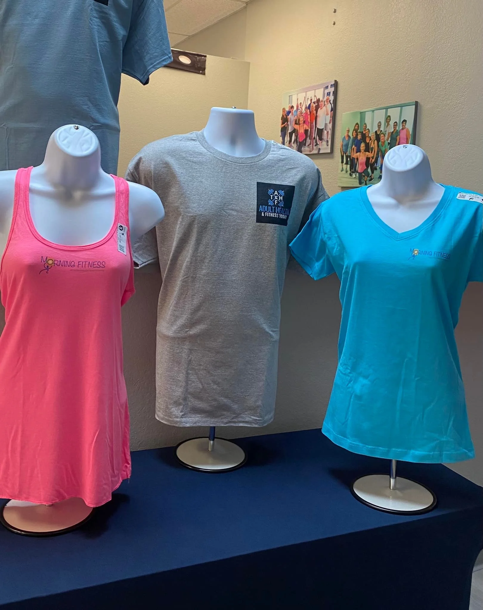 Three mannequins dressed in athletic shirts on a blue surface, with two framed photos of groups of people exercising on the wall behind them.