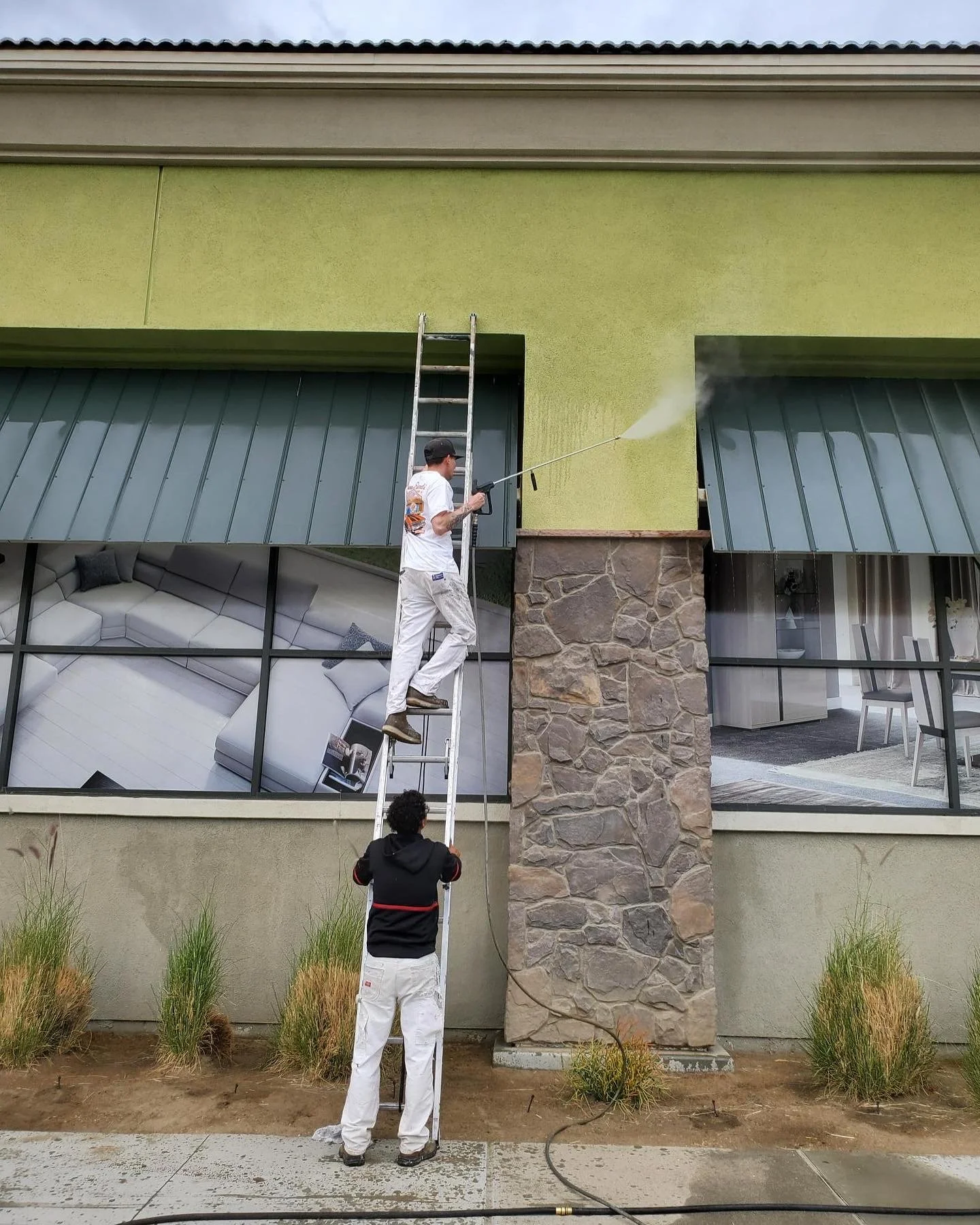 Two workers are cleaning the large exterior window of a building, one standing on a ladder and the other holding the ladder at ground level.
