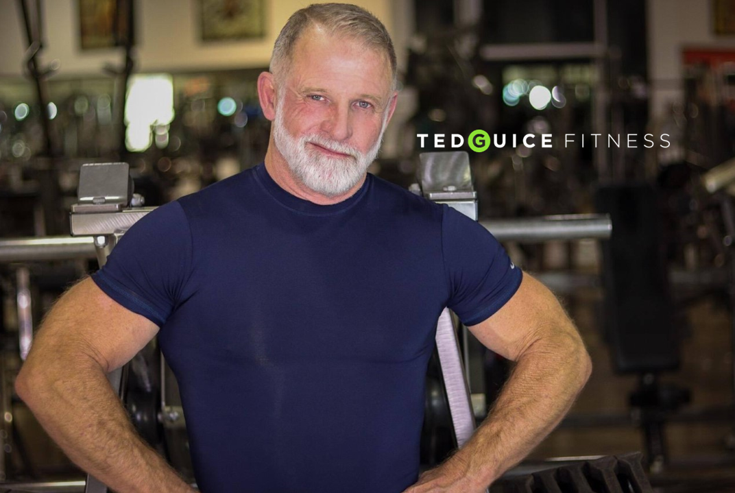 A middle-aged man with a beard and gray hair, wearing a navy blue T-shirt, standing in a gym with fitness equipment in the background. The text 'TED GUICE FITNESS' is displayed on the image.