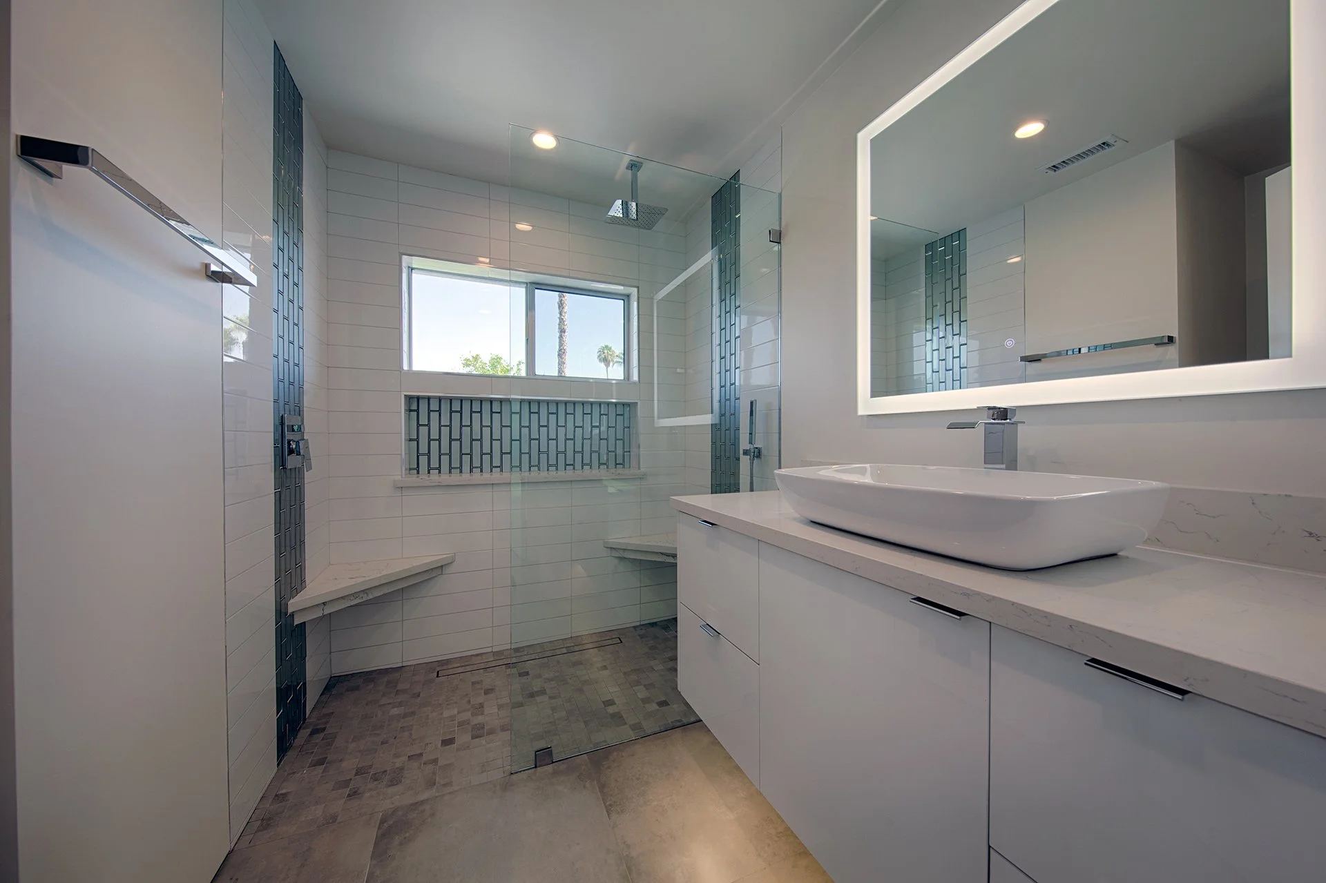 Modern bathroom with a large rectangular mirror, vessel sink on a white countertop, glass-enclosed shower with a window, white wall tiles with vertical blue tile accents, and wood-like tile flooring.
