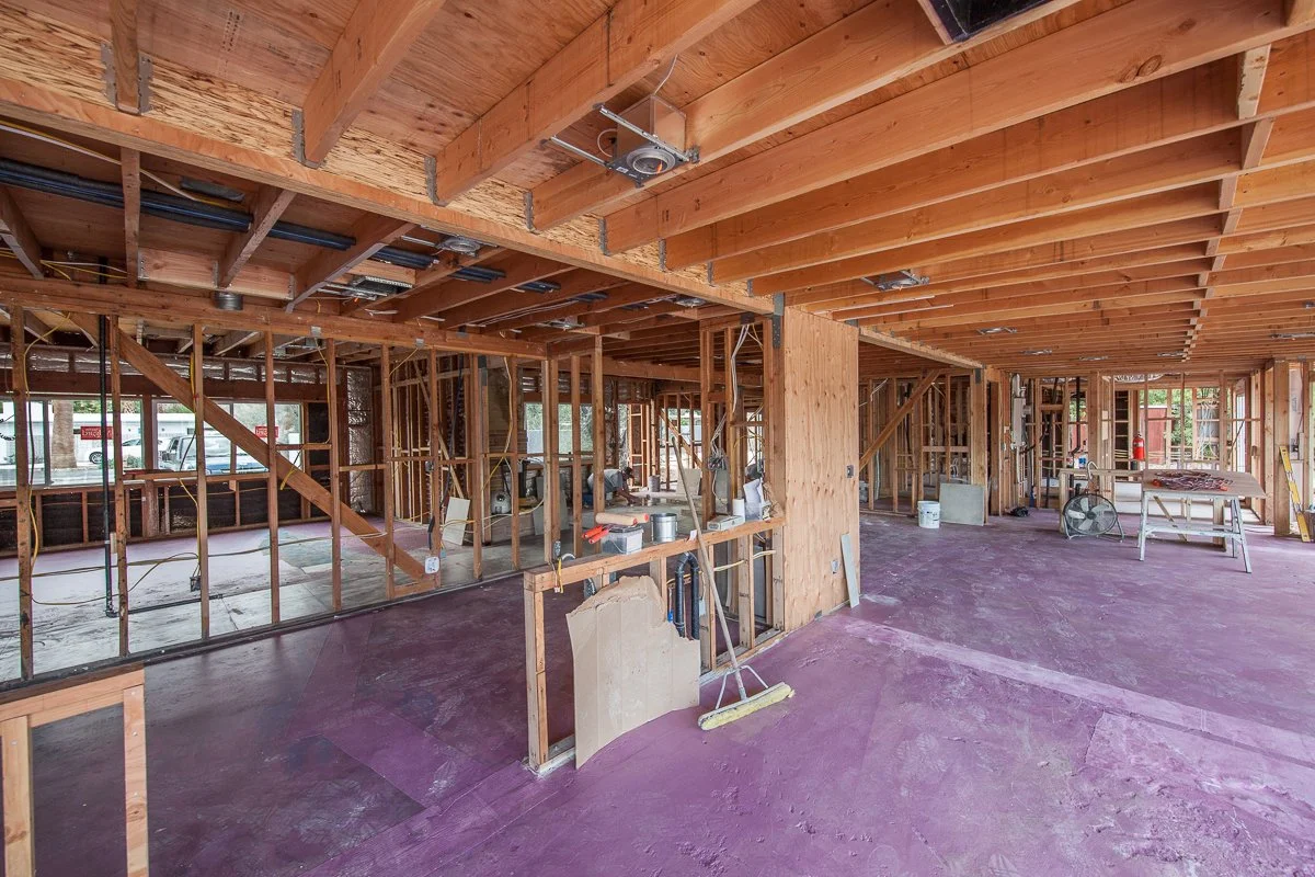 Interior view of a building under construction, showing exposed wooden framing, a partially finished floor, and construction tools and materials scattered around.