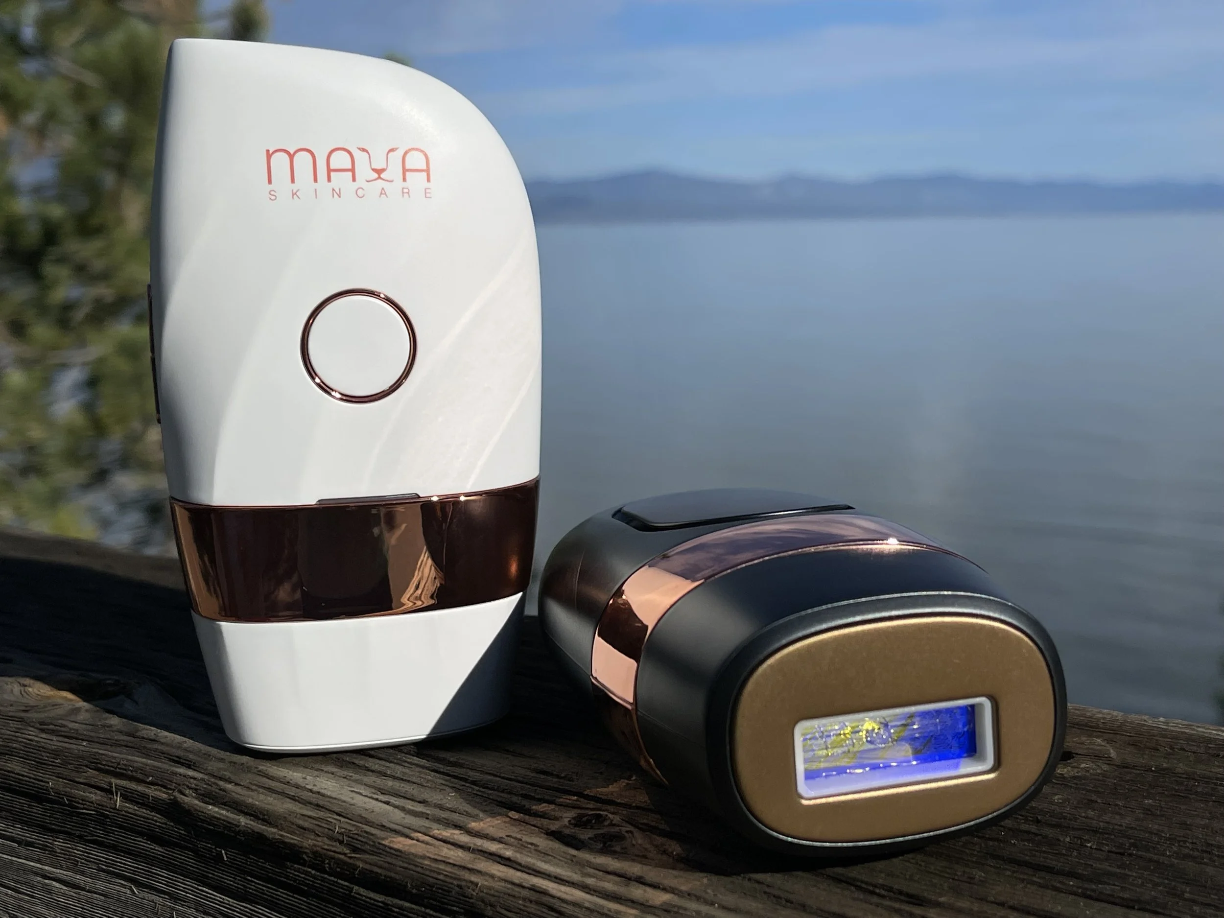 A white MAXA skincare device with a round copper ring and a black base, along with a smaller black and copper device, placed on a wooden surface outdoors near water with a blurred background of trees and mountains.