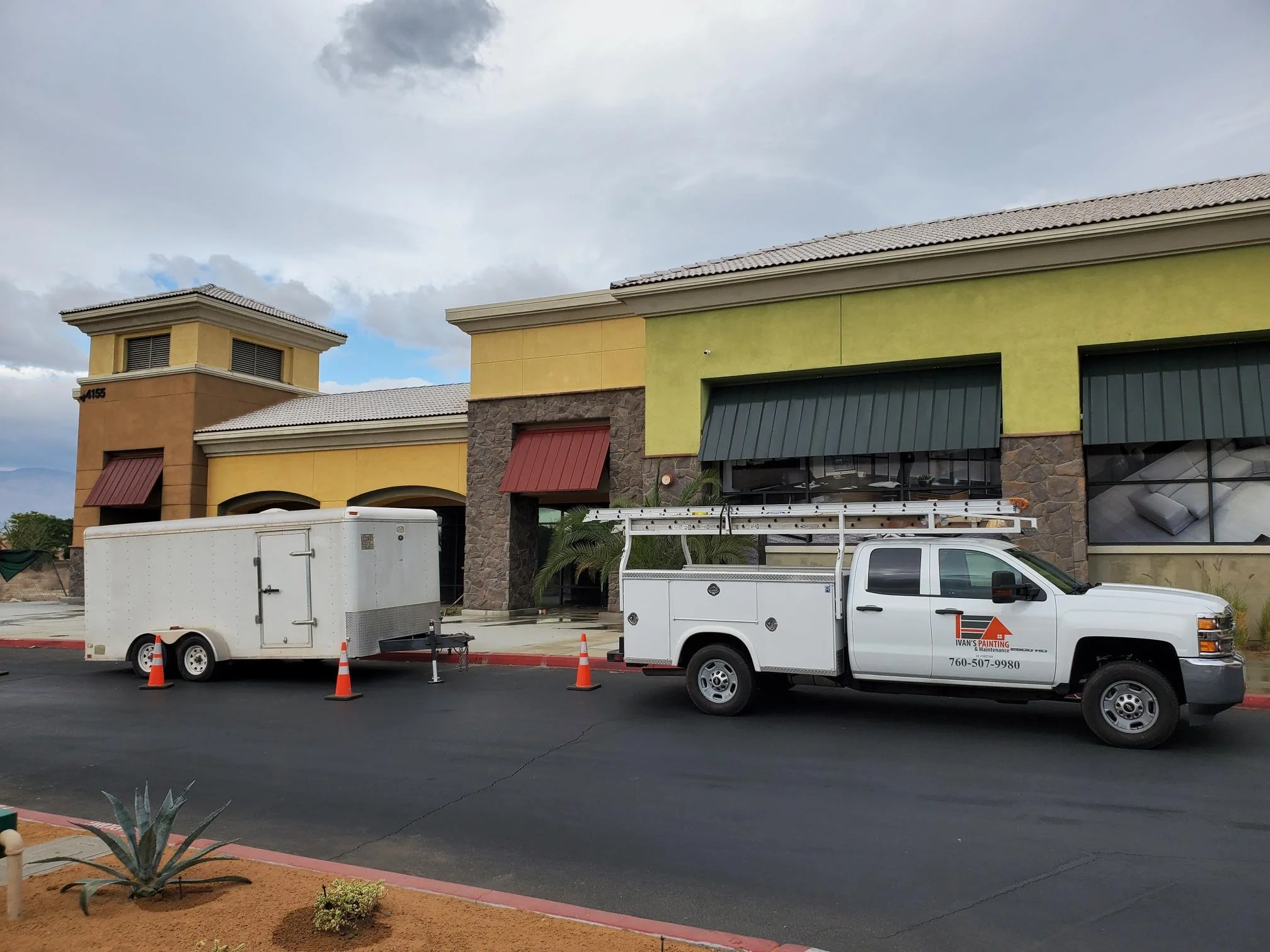 Front view of commercial building with green and beige exterior walls, red and green awnings, and a tower structure. Two white utility trucks parked in front, with orange traffic cones around. Sky is cloudy.