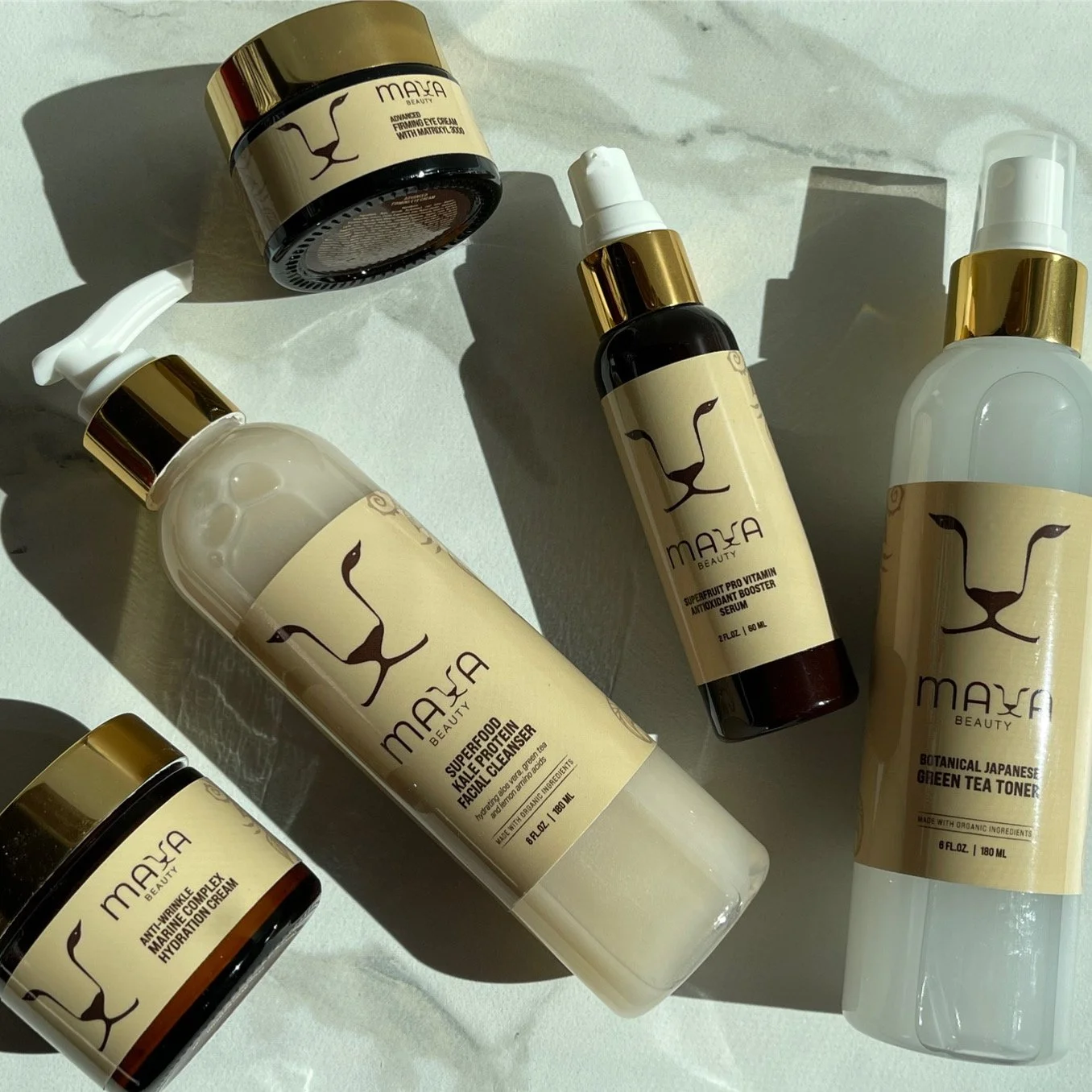 Collection of skincare products from Maya Beauty including facial cleanser, vitamin serum, green tea toner, and hydrating cream, arranged on a light surface with shadows.