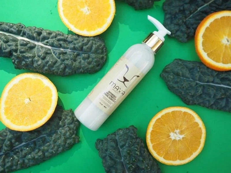 A bottle of skincare product surrounded by halved oranges and black kale leaves on a green background.