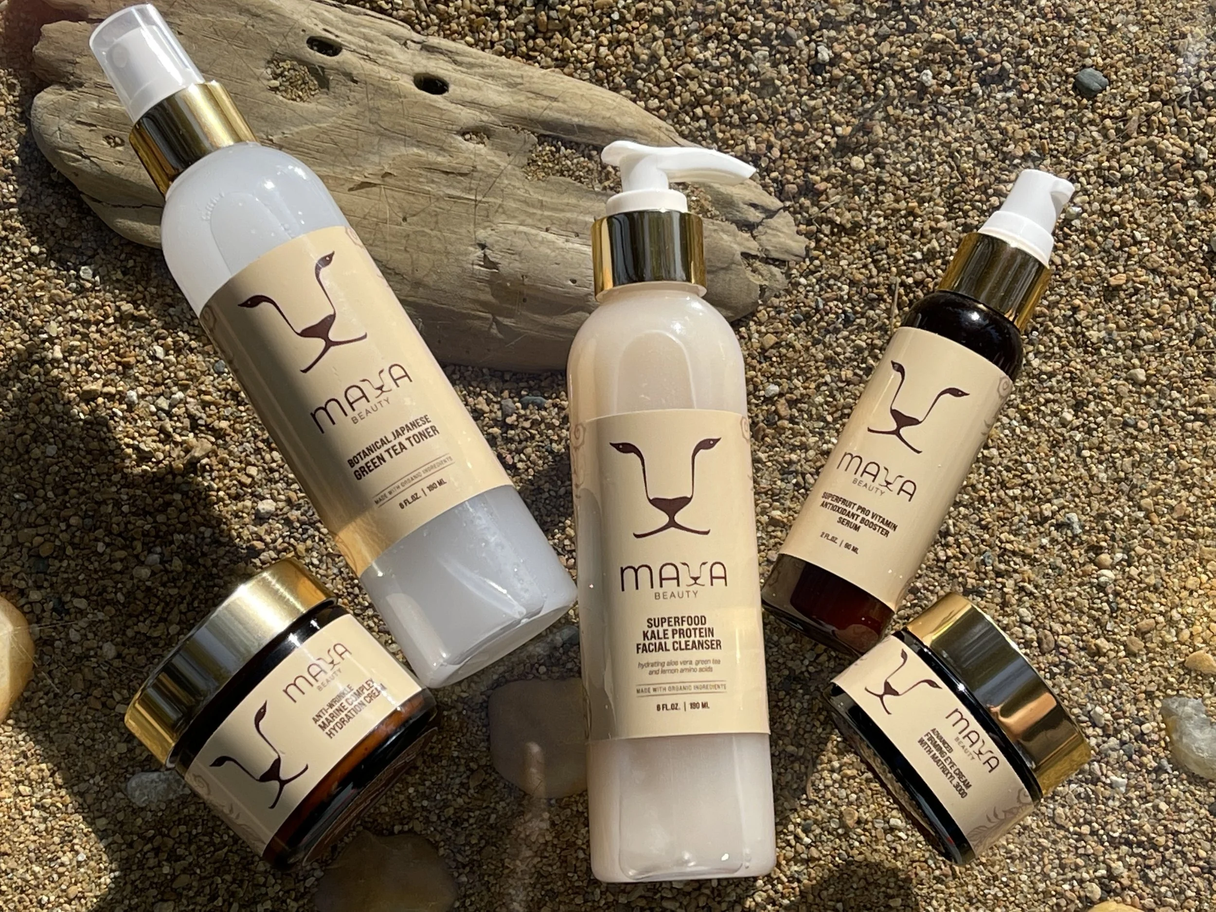 Multiple skincare bottles with gold caps and labels showing a lion's face logo, lying on a sandy beach with small pebbles and a piece of driftwood.