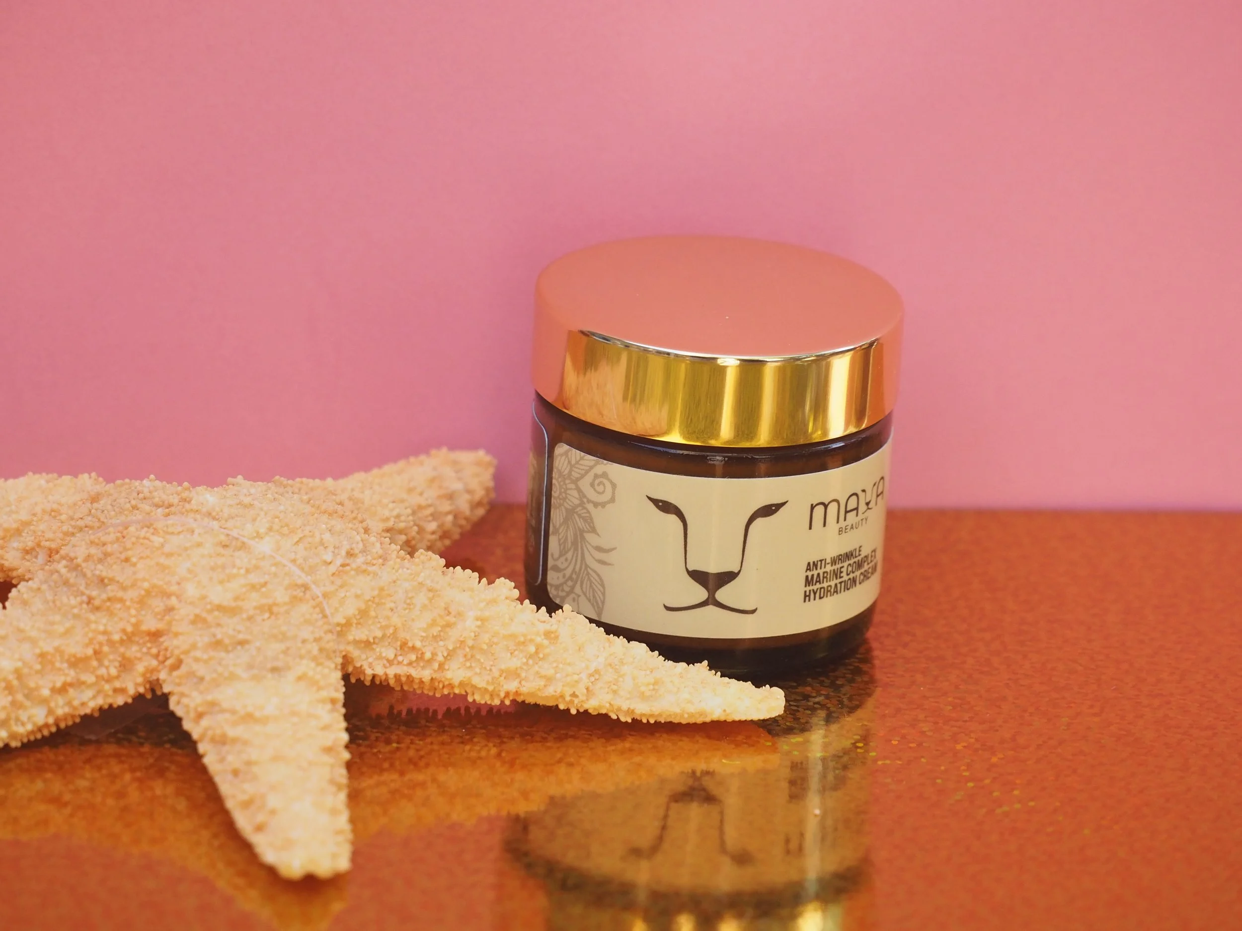 A small jar of Mayan Beauty anti-wrinkle marine collagen hydration cream with a gold lid, placed on a brown surface beside a flat, round bread or cracker on a wooden table with a pink background.