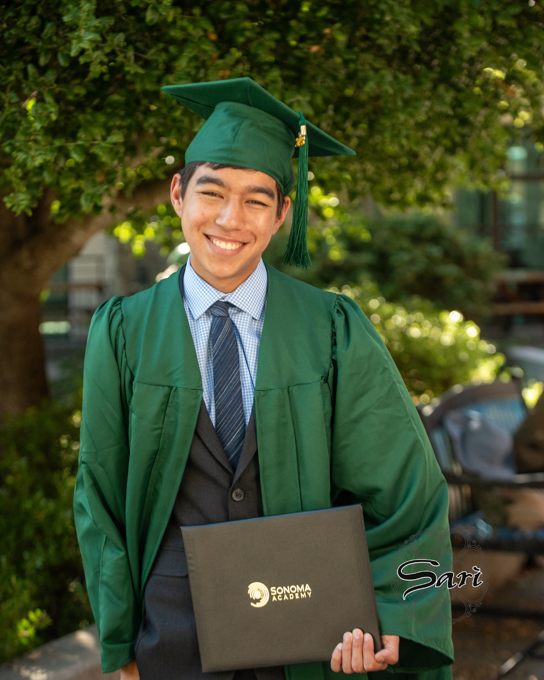 A young man wearing a green graduation cap and gown holding a black diploma cover, smiling outdoors with trees and benches in the background.