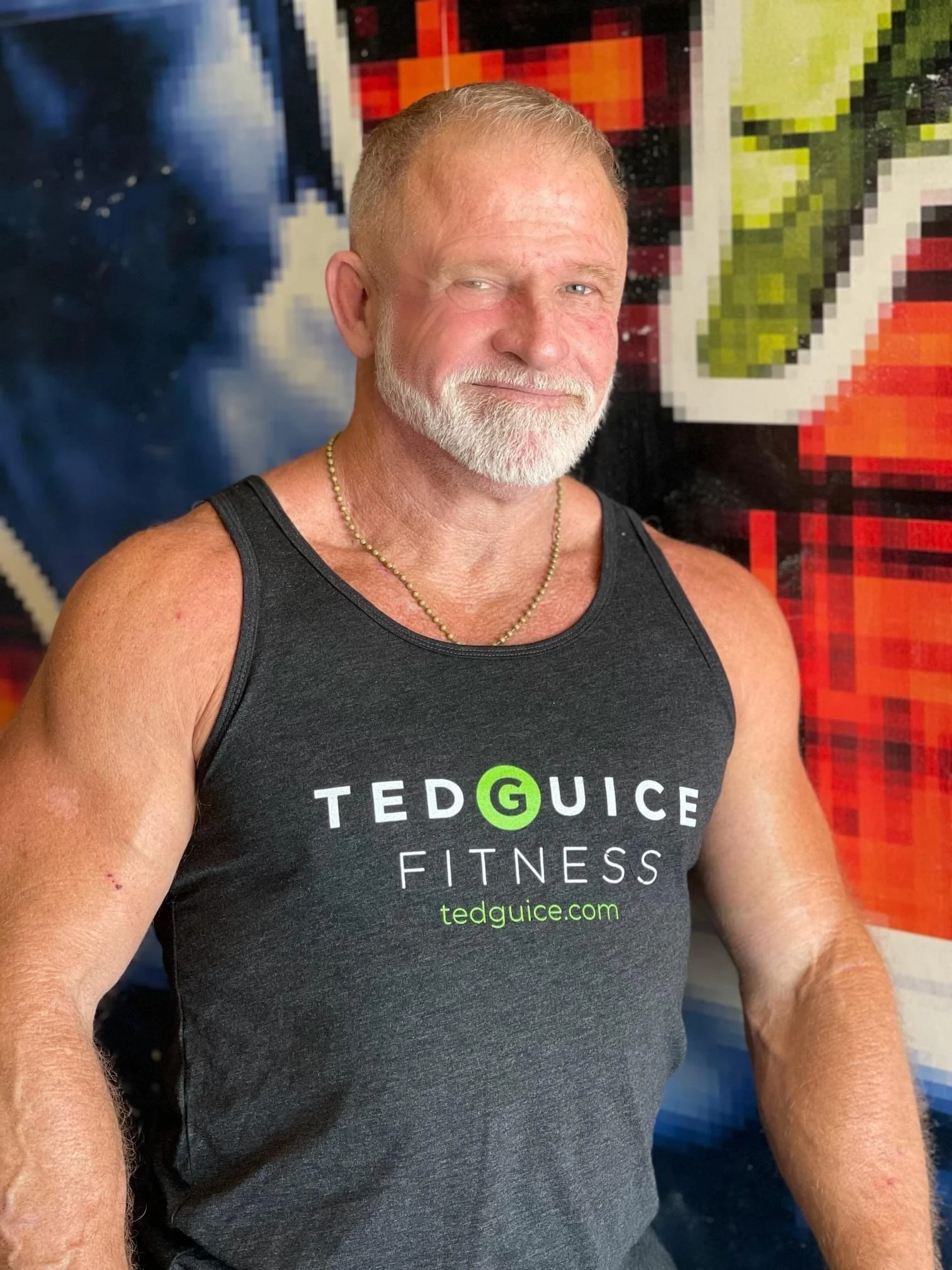 A muscular man with a white beard and short hair taking a selfie, wearing a black tank top with 'Ted Guice Fitness' written on it, standing in front of a colorful, pixelated background.