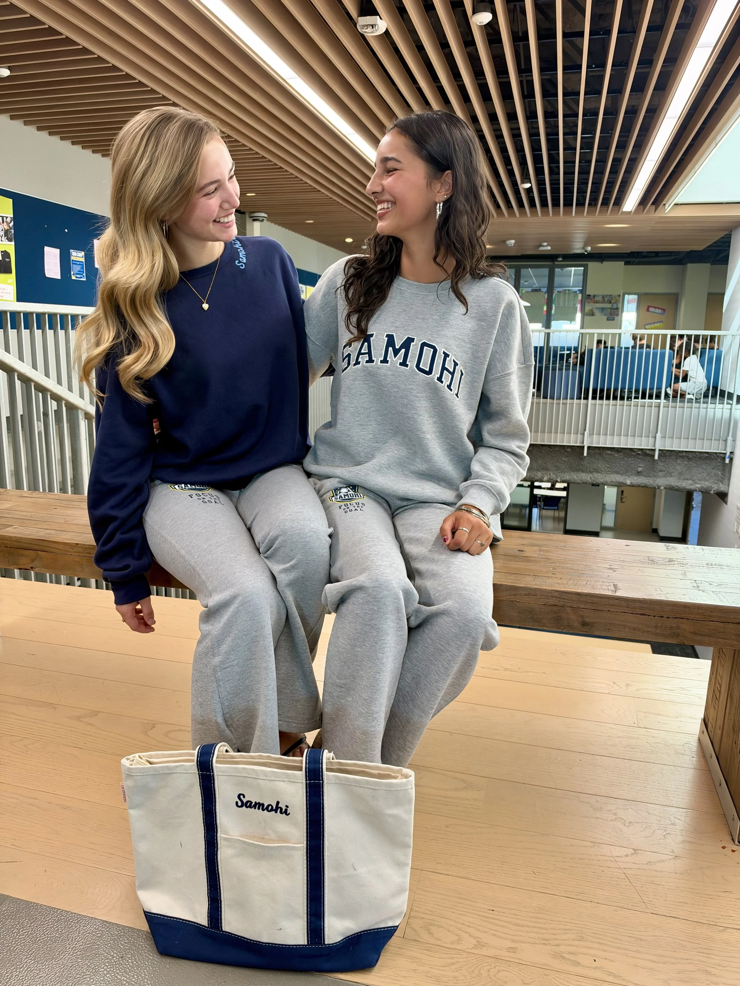 Two smiling women in Samohi sweatshirts sitting on wooden bleachers inside a school, with a Samohi tote bag in the foreground.