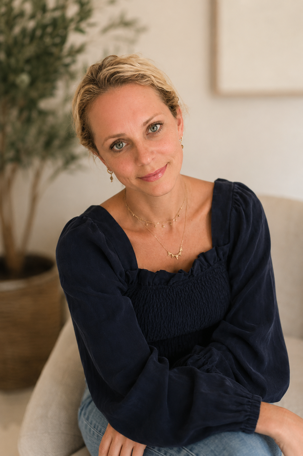 A woman with blonde hair and blue eyes, wearing a navy blue long-sleeve top and layered gold jewelry, sits on a beige chair in a softly lit room with a potted plant in the background.