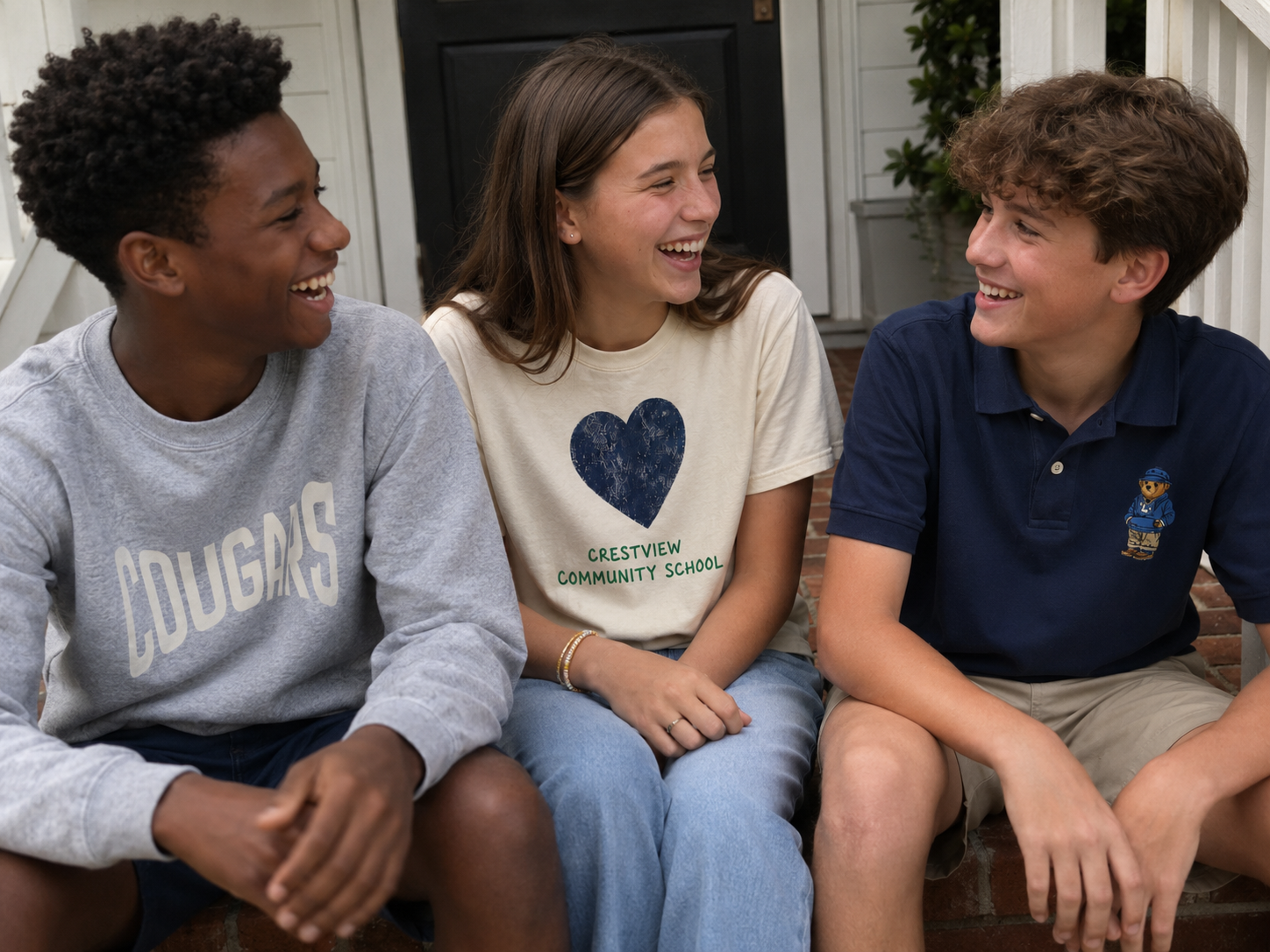 Three teenagers sitting on steps outside, smiling and laughing together, with a girl in the middle wearing a Crestview Community School t-shirt.