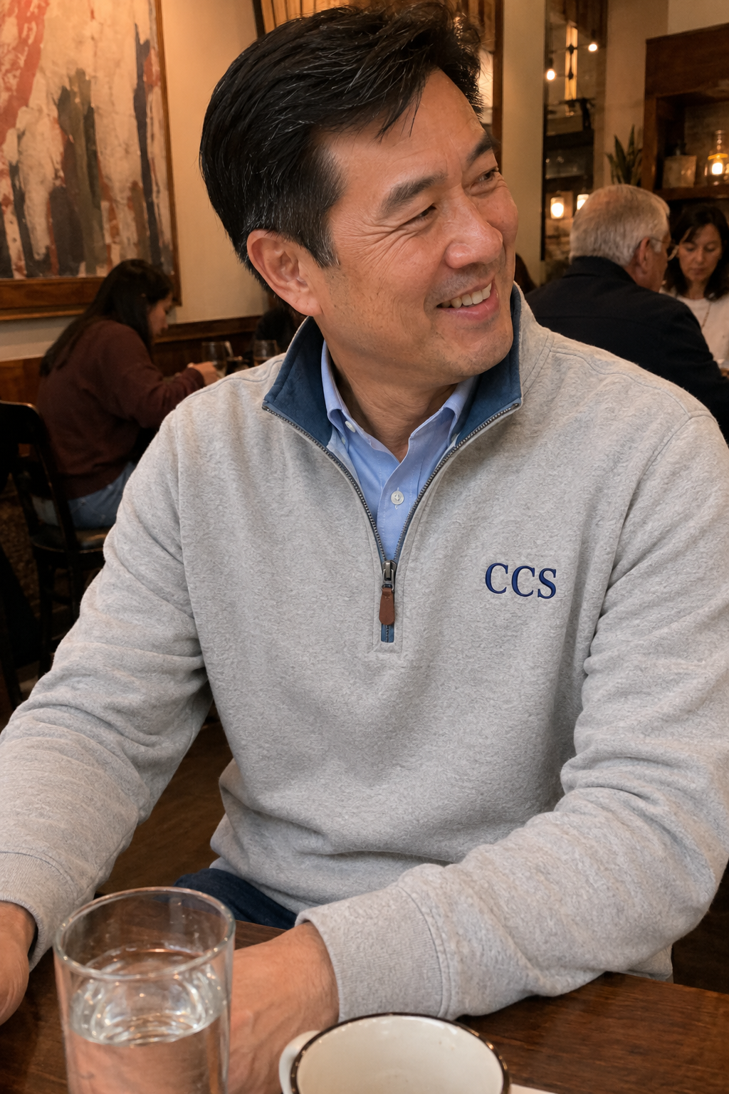 A smiling middle-aged man with black hair, wearing a gray sweatshirt with 'CCS' embroidered on it, and a light blue collared shirt underneath, sitting at a wooden table in a restaurant, with a glass of water and a cup nearby.