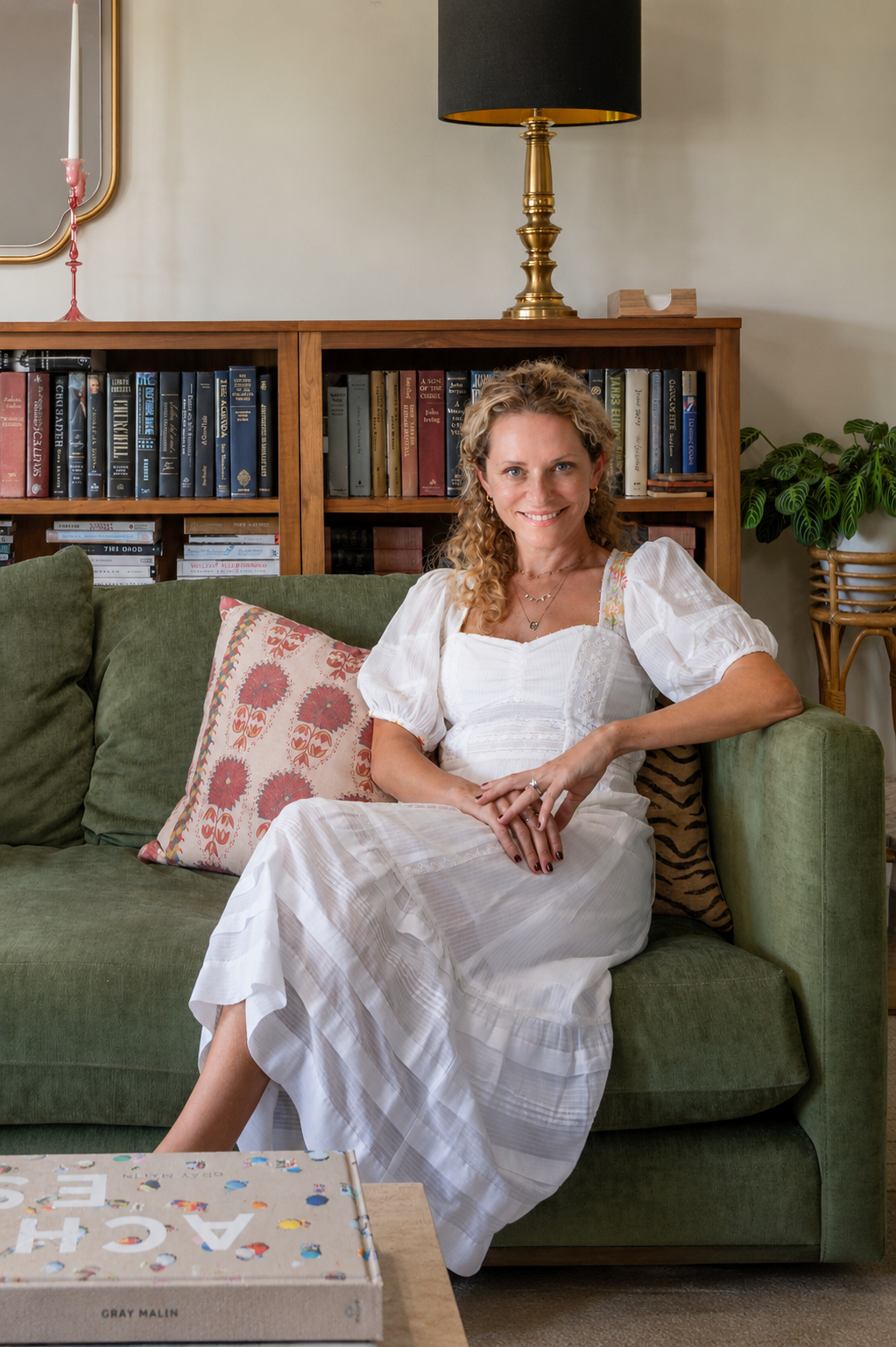 A woman sitting on a green sofa, smiling, with a book in front and a wooden bookshelf behind her filled with books. There's a gold and black table lamp on top of the bookshelf and a pink candlestick on the left side. A decorative pillow with floral patterns is on the sofa and a green potted plant is on the right side.