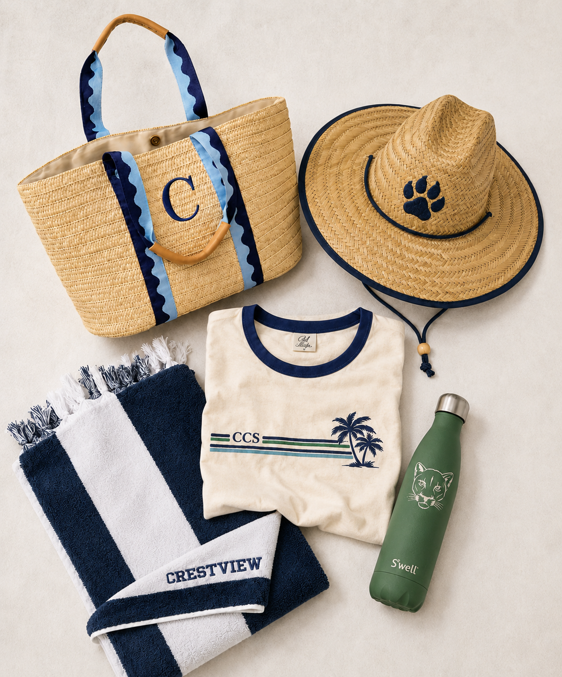 A beige straw beach bag with blue accents and a monogrammed initial, a matching straw sunhat with a paw print, a cream T-shirt with a palm tree design and the letters CCS, a navy and white striped towel with a Crestview logo, a green water bottle with a panther face, and a white and navy towel with fringes.