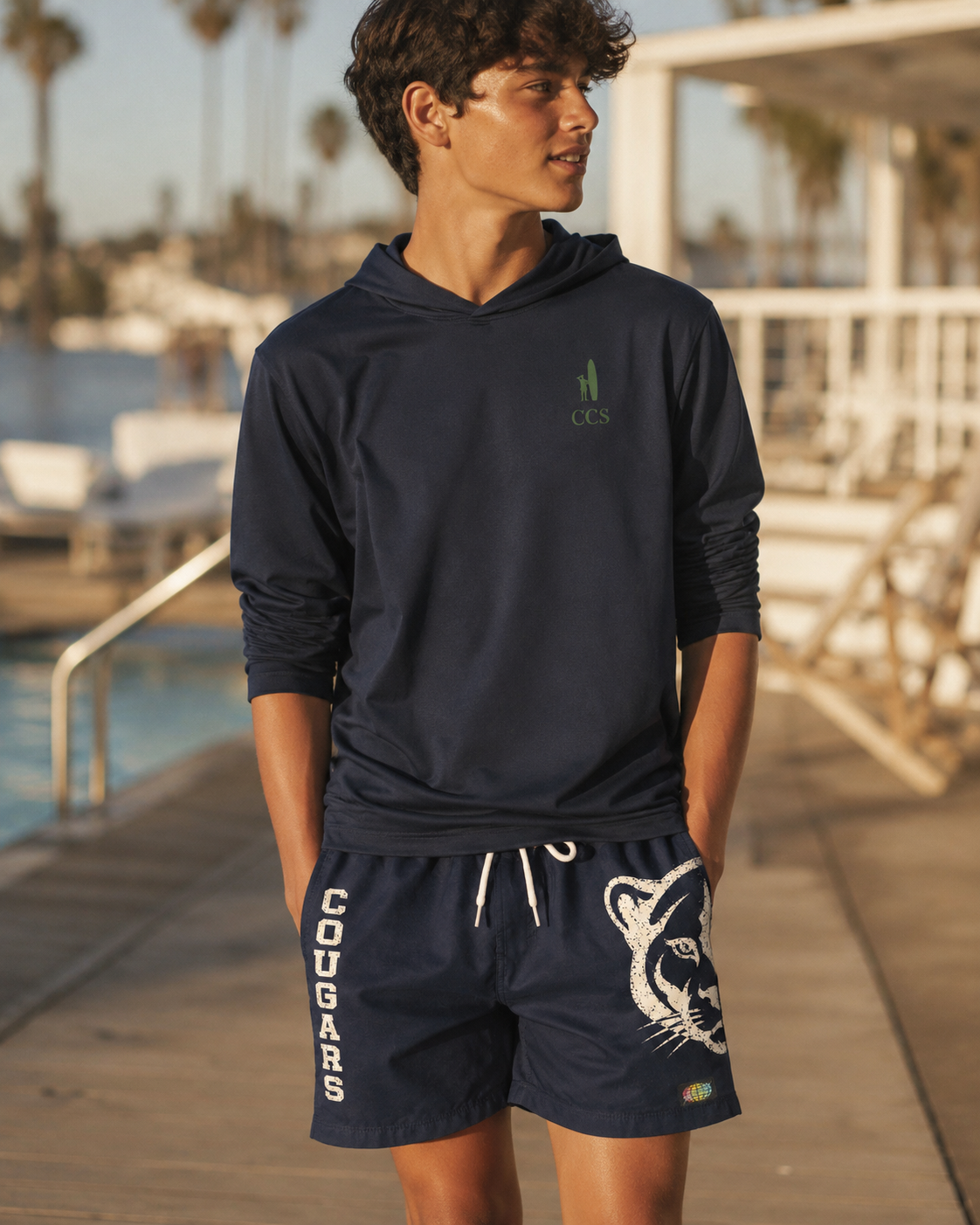 A young man standing on a wooden dock near the water, wearing a dark hooded athletic shirt and shorts with a cougar print and the word "COUGARS" on them. He has his hands in his pockets and is looking to the side.