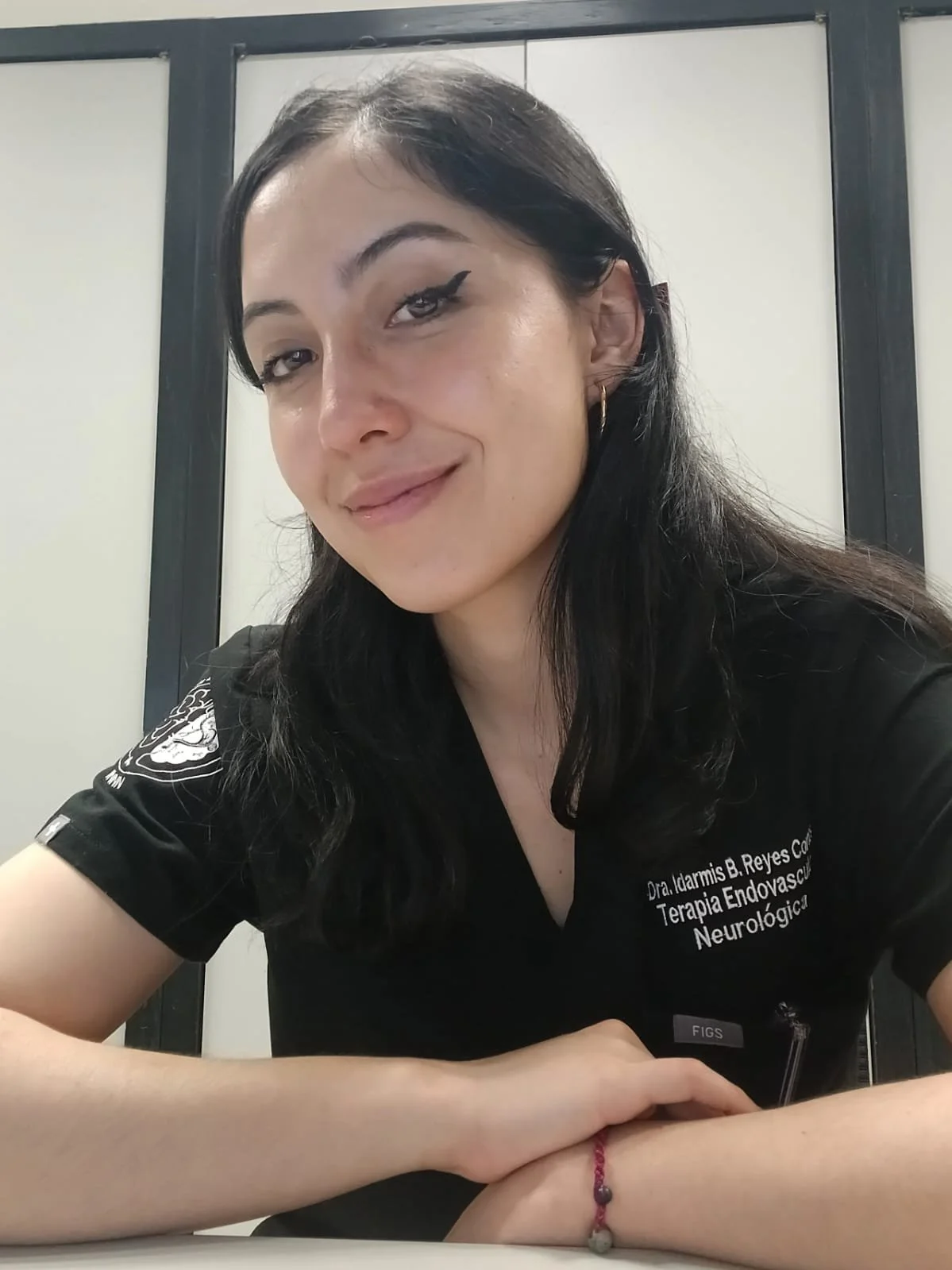 Mujer con cabello largo y oscuro, usando una camiseta negra con texto en español, en un fondo de paredes blancas y marco negro.