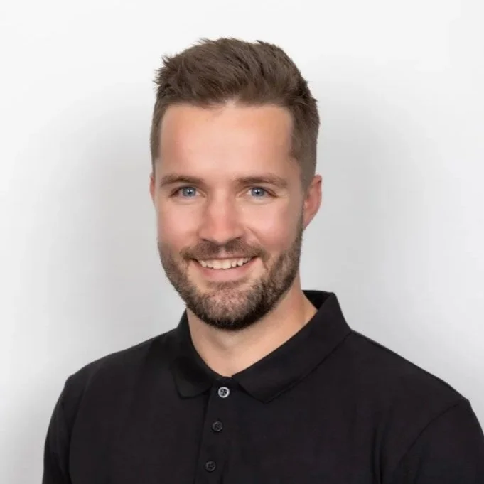 A portrait of a smiling man with short brown hair, beard, and blue eyes, wearing a black collared shirt against a plain white background.